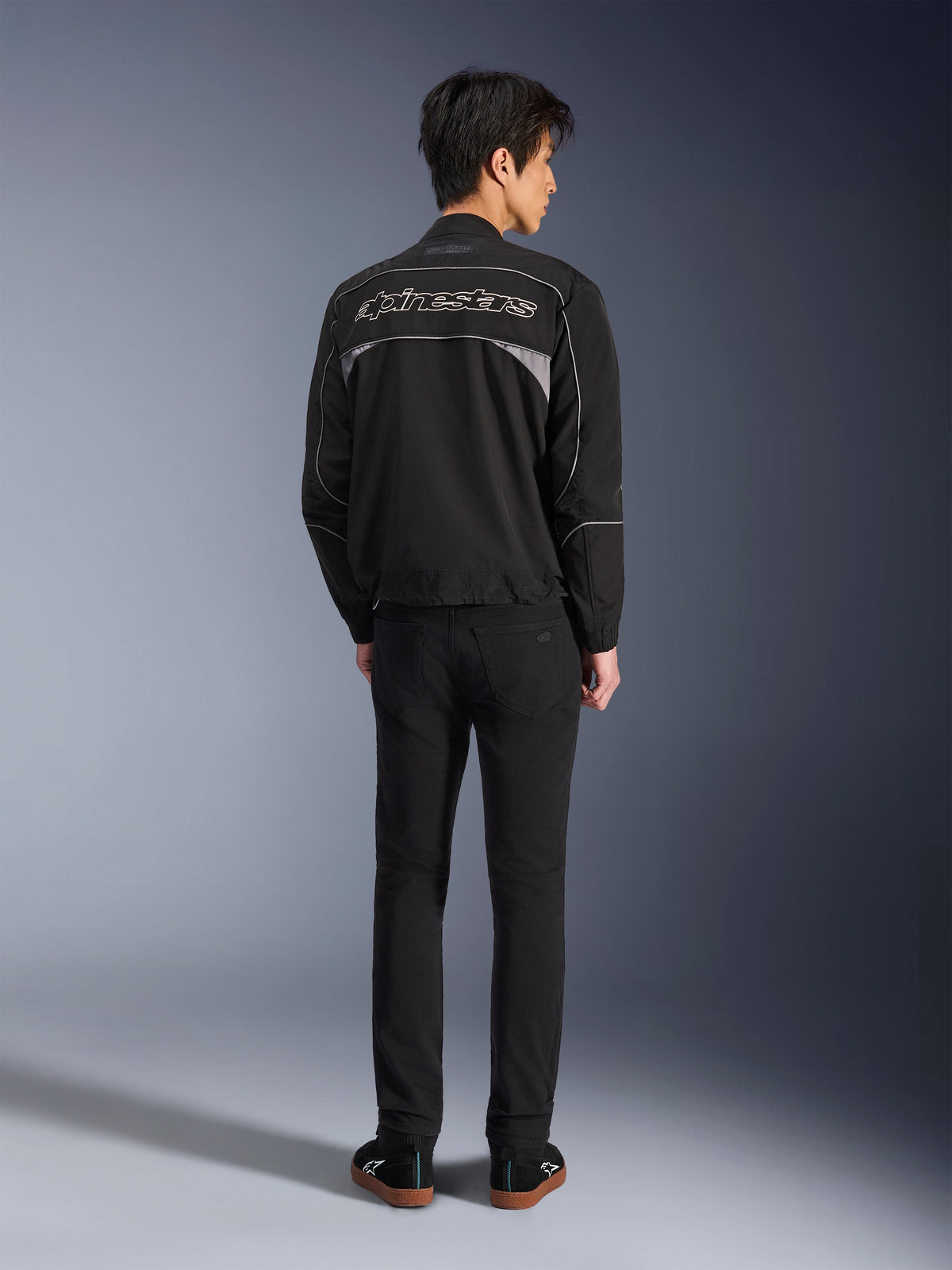 Alpinestars Unite Jacket, Black and Dark Gray motorcycle jacket, rear view showing Alpinestars outline logo on the back, gray reflective panels, minimalist urban design, worn with black pants and black shoes.