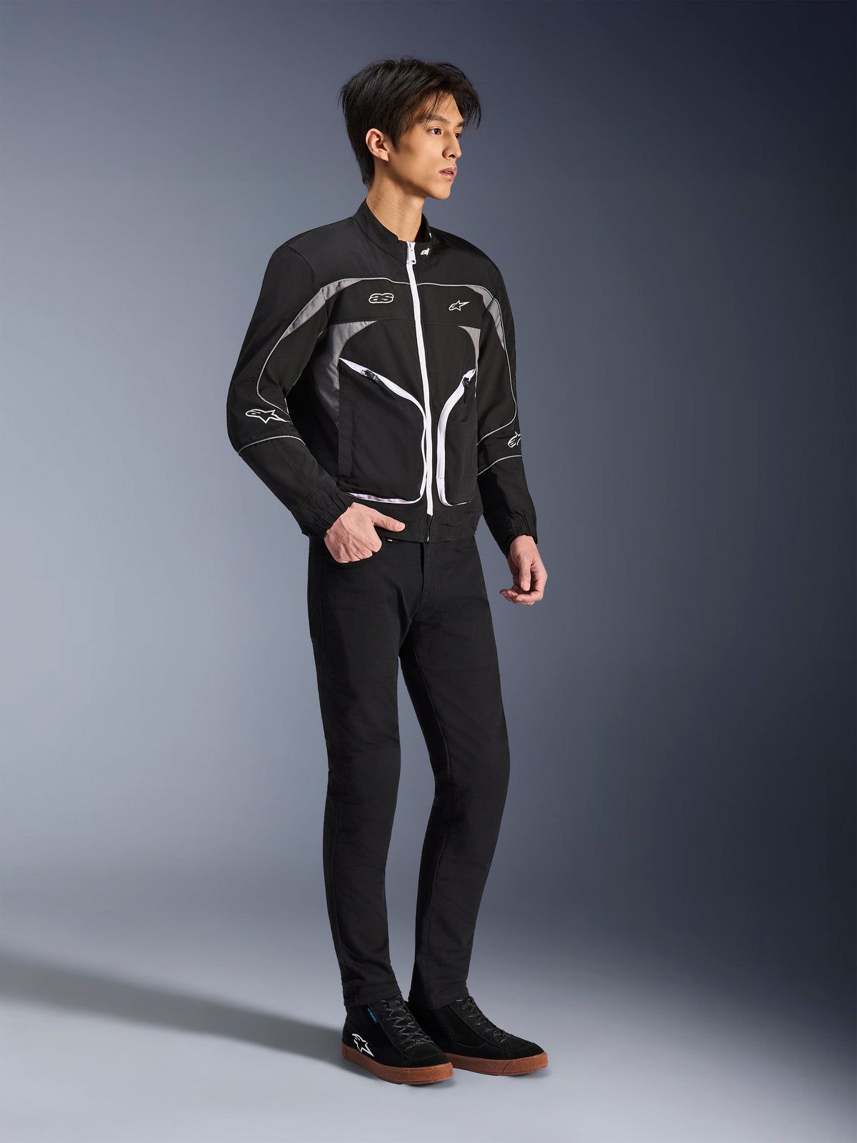 Model wearing Alpinestars Unite Jacket in Black and Dark Gray, Urban Motorcycle Jacket featuring white zippers and reflective details, paired with black slim-fit pants and sneakers, minimalist street-inspired riding gear.