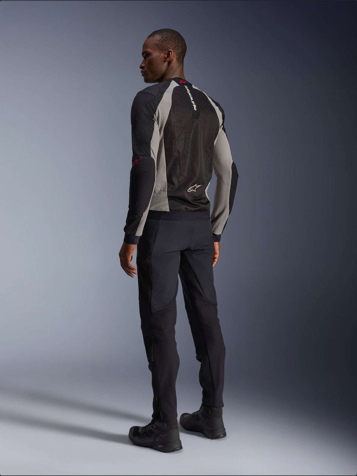Model wearing Alpinestars Troop-Air Jacket from the back, Black Ash Gray and Bright Red accents, technical mesh construction for ventilation, paired with black motorcycle pants and riding boots, urban contemporary style