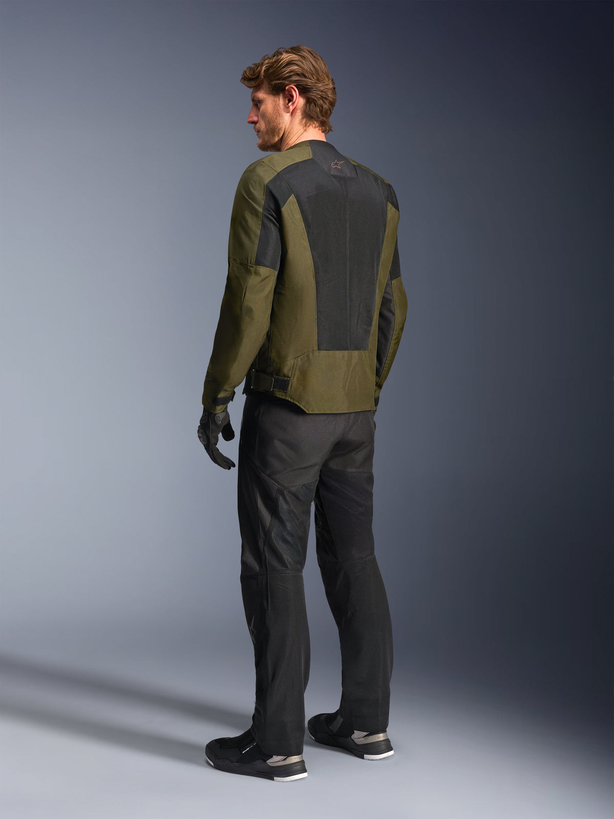 Male model wearing Alpinestars C-1 Air Jacket in Khaki, urban motorcycle jacket, rear view highlighting large black mesh panels on the back, paired with black technical riding pants and gloves, contemporary motorcycle gear for warm weather