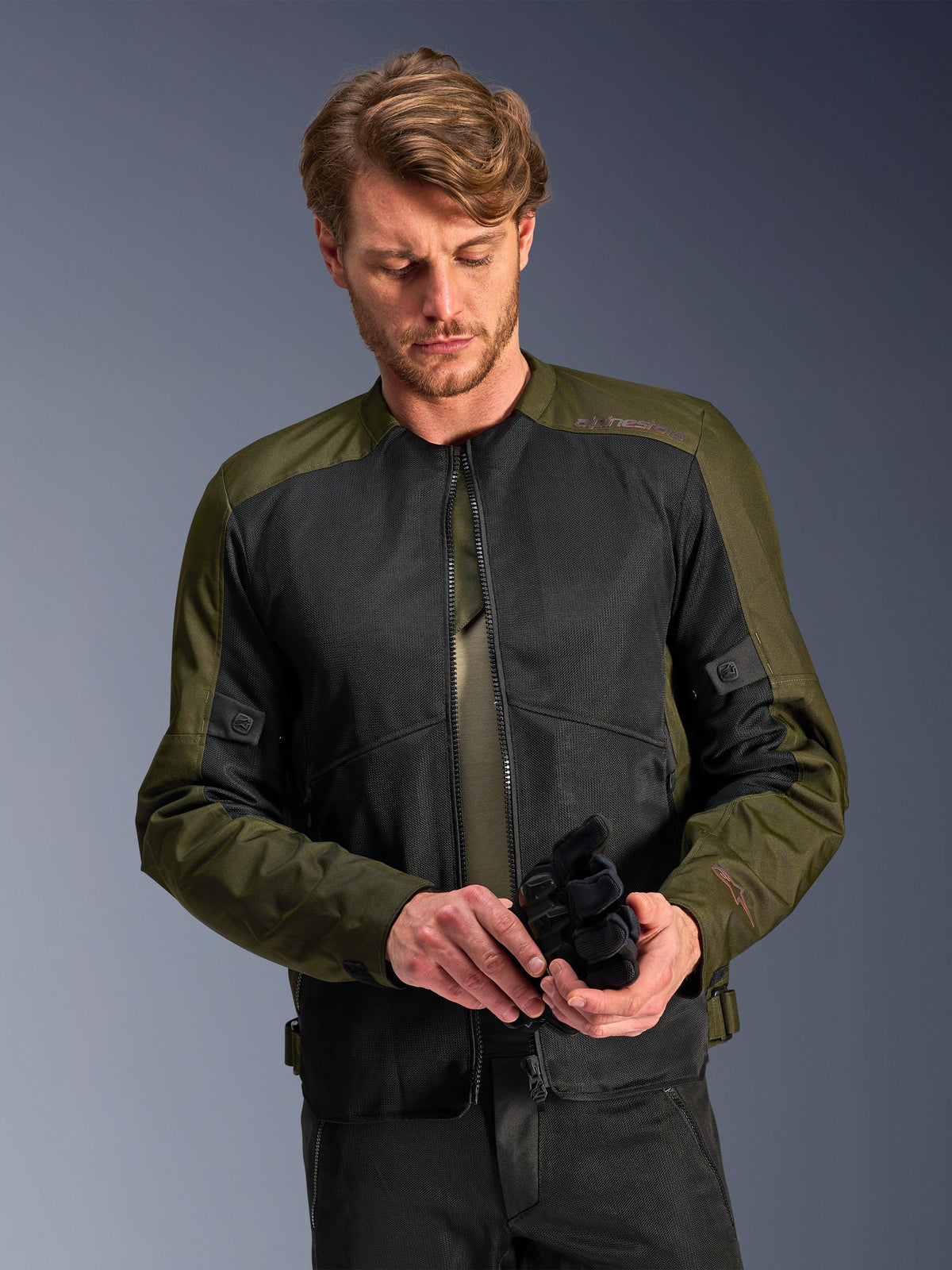 Man wearing the Alpinestars C-1 Air Jacket in Khaki, urban motorcycle jacket with olive green textile and black mesh panels, looking down while holding black motorcycle gloves, clean and contemporary style for road riding.