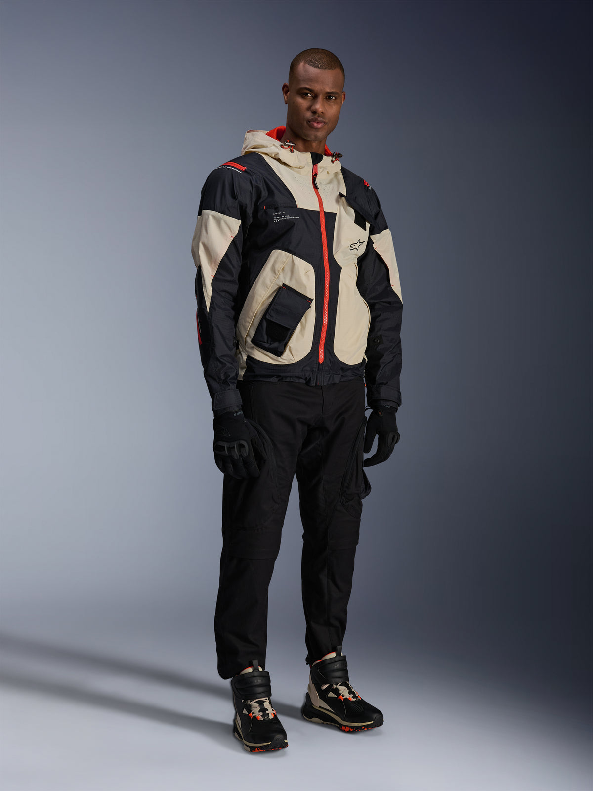 Model wearing the Alpinestars Mogress Air Jacket in Black, Almond, and Hot Orange, urban motorcycle jacket with a utility-inspired design, worn with black riding pants, black gloves, and coordinated technical boots, contemporary street-ready riding gear
