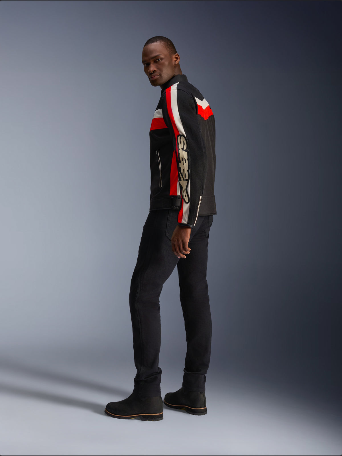 A man wearing the Alpinestars T-Dyno Air Jacket in Black, Bright Red, and Ecru, vintage-inspired textile motorcycle jacket with mesh panels, bold branding on the sleeve, paired with black pants and boots for a modern biker look
