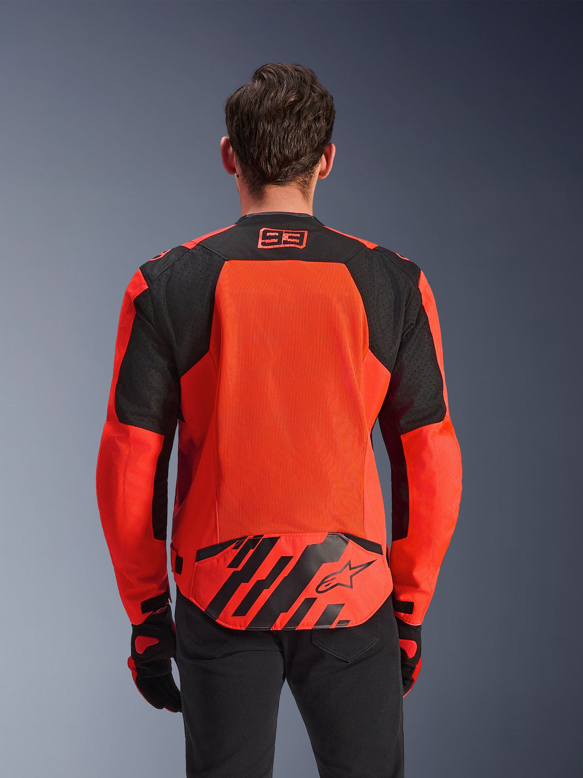 Alpinestars MM93 T-SPS Air V2 Jacket, Rear View, Red Fluo and Black, featuring 93 racing logo on the neck, large mesh panel on the back for airflow, lower back flap with Alpinestars star logo and geometric stripes, worn with black pants and riding gloves