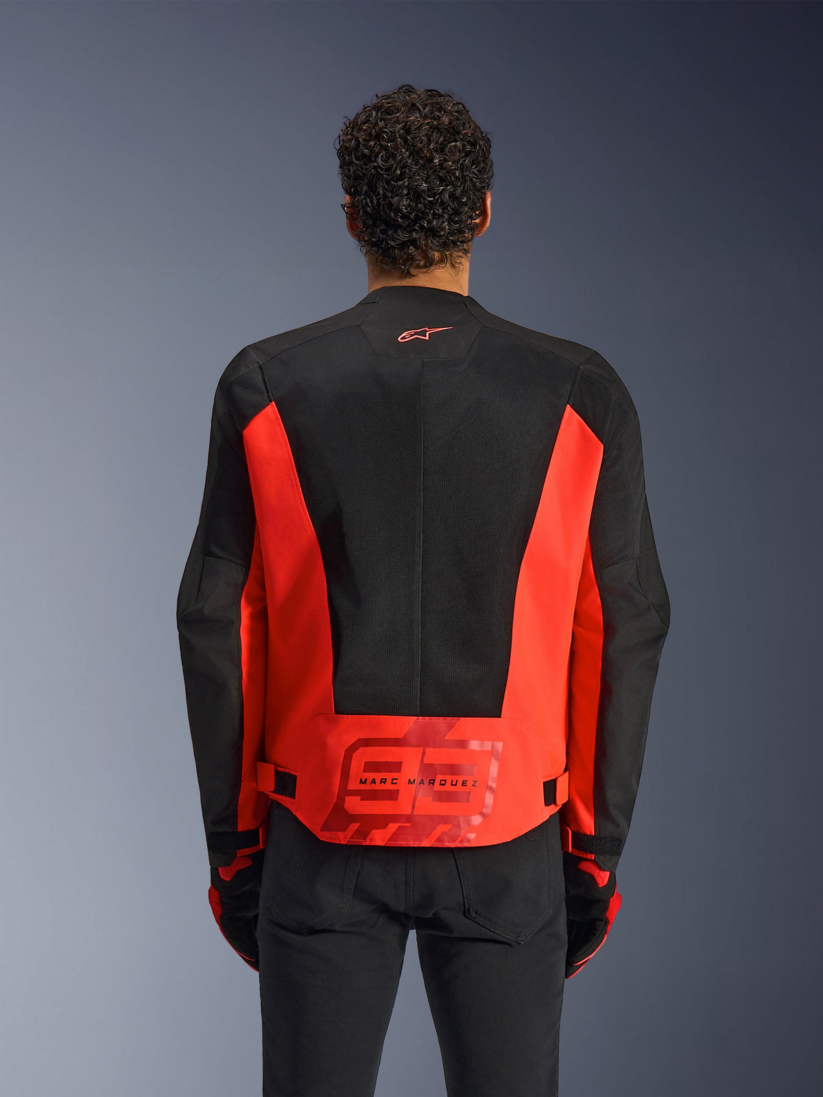 Rear view of a man wearing the Alpinestars MM93 C-1 Air Jacket in Red Fluo and Black, featuring a large '93' Marc Marquez logo on the lower back, lightweight mesh panels for ventilation, paired with black pants and gloves.