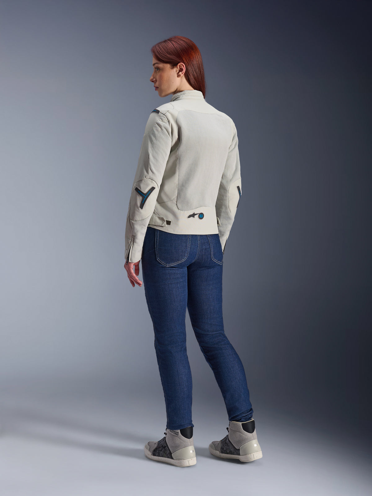 Woman wearing Alpinestars Stella Circal-1 Air Jacket in Metal Gray and Methyl Blue, rear view showcasing mesh airflow panels and neon blue accents, paired with dark blue riding jeans and gray sneakers, urban motorcycle lifestyle shot.