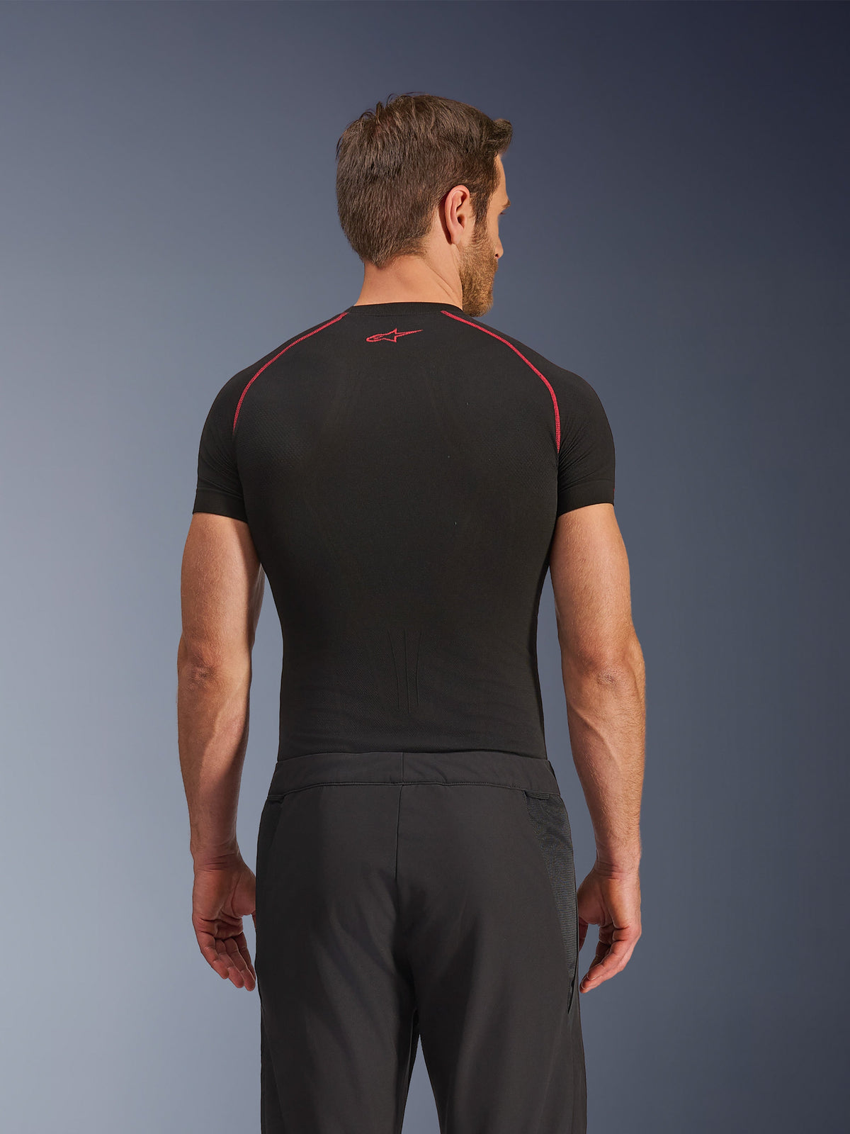 Rear view of a model wearing Alpinestars Aeroshell Airflow Pants, Black, lightweight softshell construction with integrated mesh side panels, paired with a black base layer featuring red contrast stitching, sporty motorcycle attire