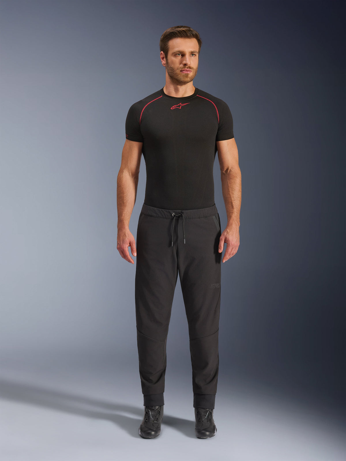 Model wearing Alpinestars Aeroshell Airflow Pants in Black, paired with a black short-sleeve base layer and black motorcycle boots. These lightweight softshell motorcycle pants feature a drawstring waist and elastic cuffs, designed for sport and touring riders in warm-weather conditions.