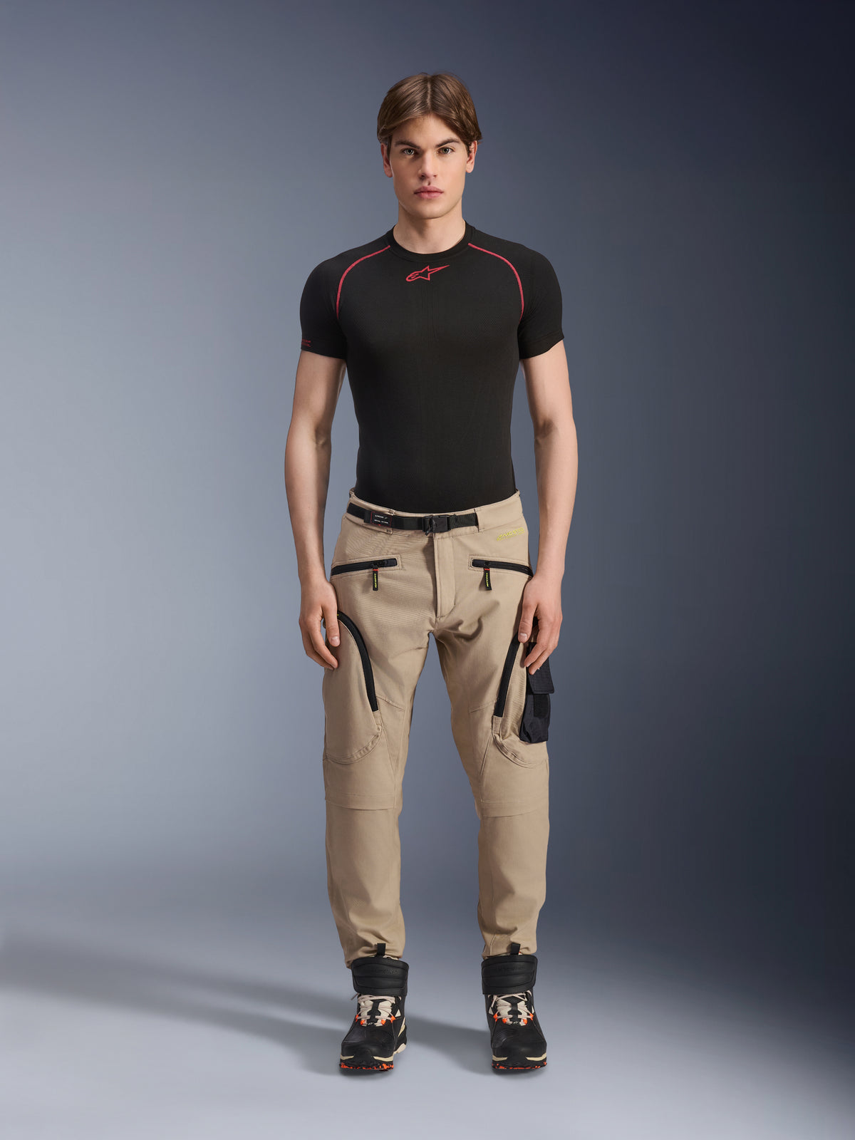 Male model wearing Alpinestars Acteon Pants in Sand, paired with a black short-sleeve base layer and black technical boots, featuring cargo pockets with black zippers and a tapered fit, contemporary urban motorcycle gear