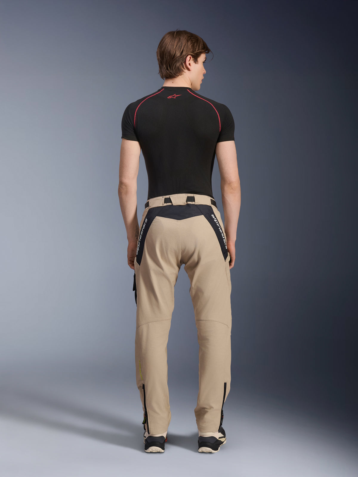 Alpinestars Acteon Pants in Sand, modern urban motorcycle pants viewed from the back, featuring black panels with white Alpinestars branding, tapered fit with zippers at the hem, worn with a black short-sleeved base layer and sneakers.