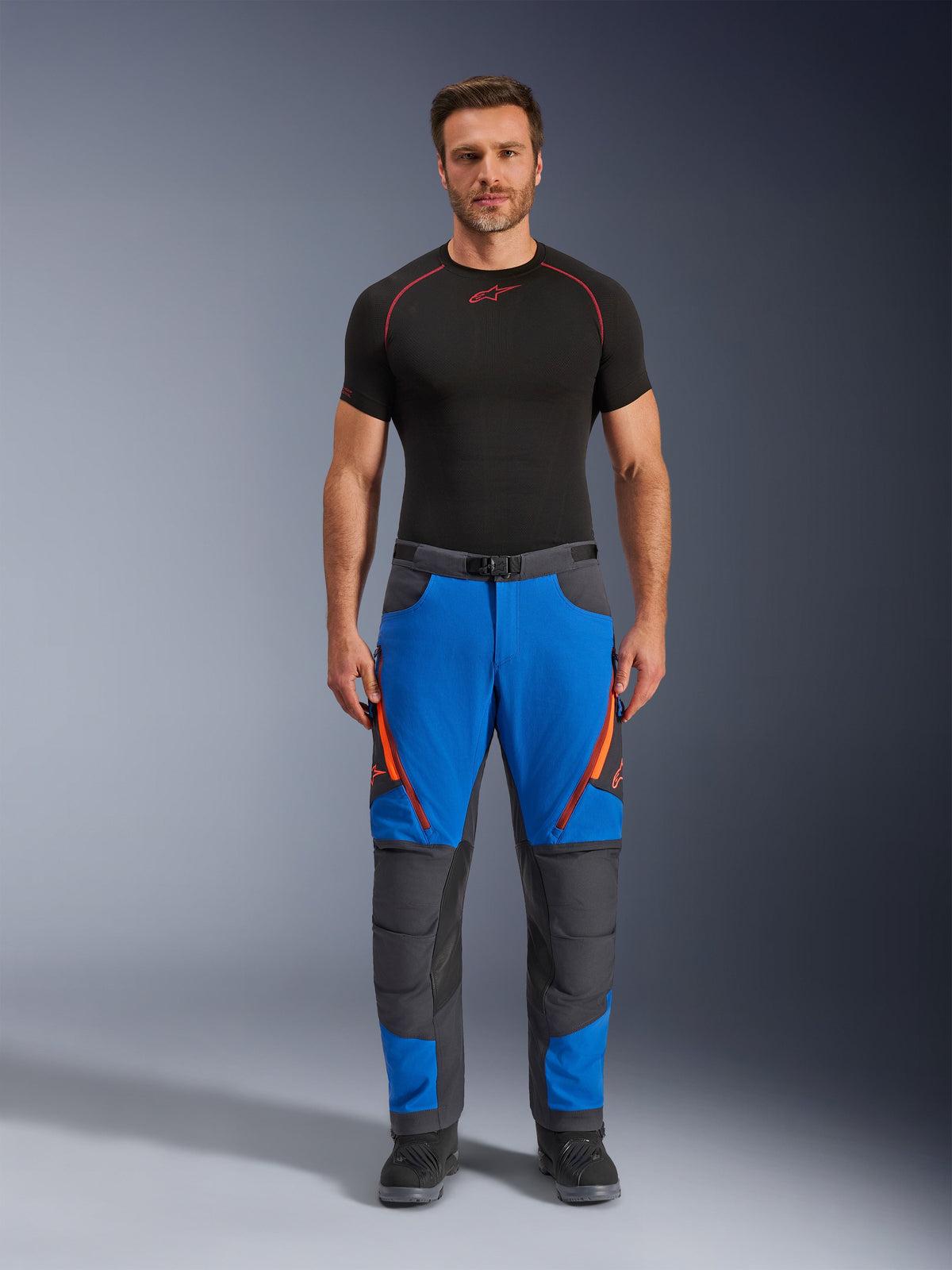 Alpinestars Maxdura Dual Pants, Adventure Motorcycle Pants, Royal Blue and Deep Gray, orange zipper accents, worn with a black Alpinestars base layer and black boots, featuring reinforced knee panels and a technical off-road design