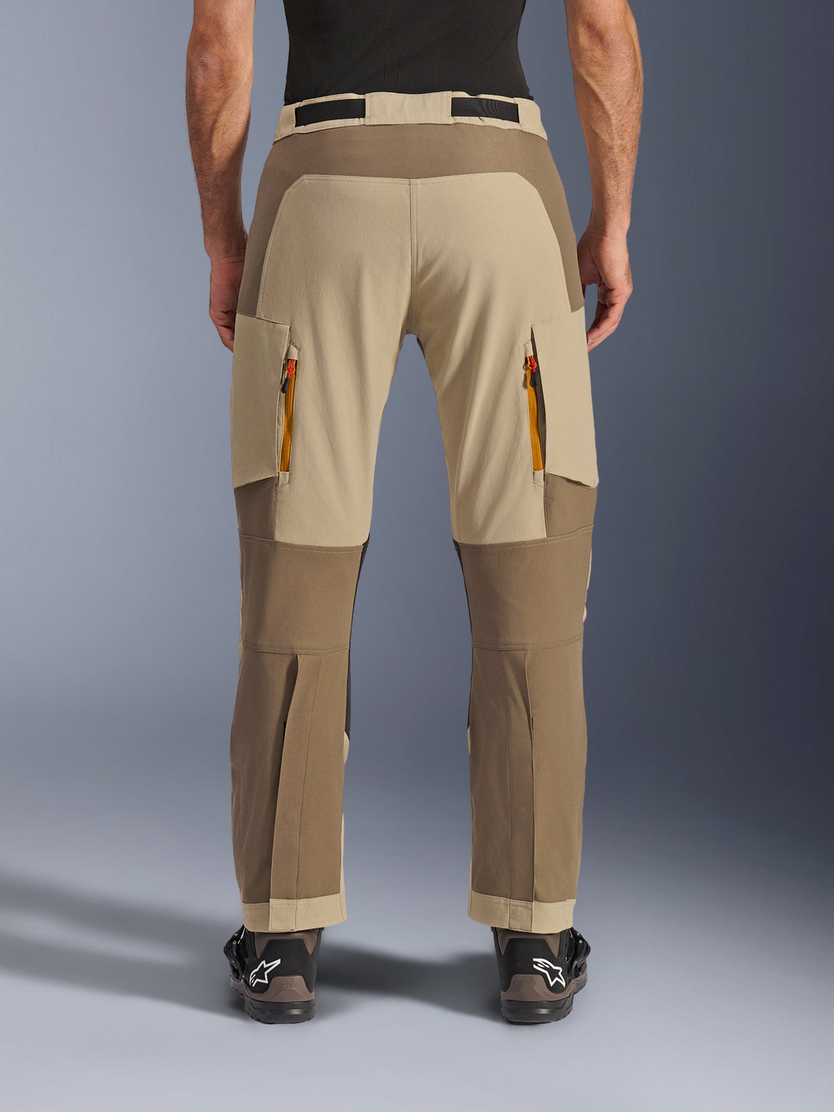 Alpinestars Maxdura Dual Pants, Walnut Aluminum, rear view of adventure motorcycle pants featuring tan and earth-tone panels, orange-zipped exhaust vents, and over-the-boot design, worn with black Alpinestars boots.