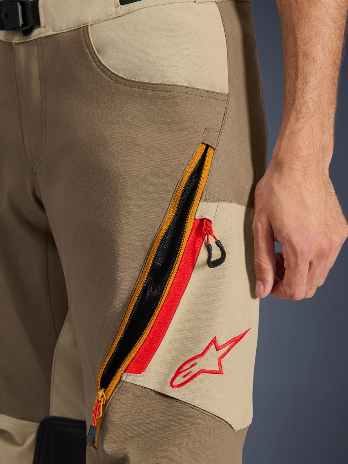 Alpinestars Maxdura Dual Pants, Walnut and Aluminum, close-up of thigh detail featuring a large diagonal zippered ventilation system with red accents and a red Alpinestars logo on a tan panel, adventure motorcycle gear