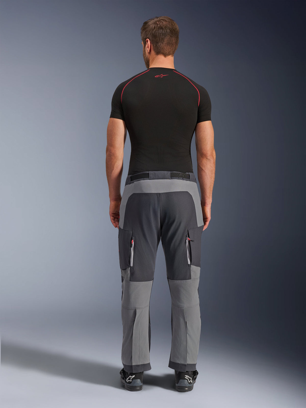Alpinestars Maxdura Dual Pants, Dark Gray and Deep Gray adventure motorcycle pants, back view on model wearing black t-shirt and boots, featuring multi-tone gray panels and zippered cargo pockets with red accents.