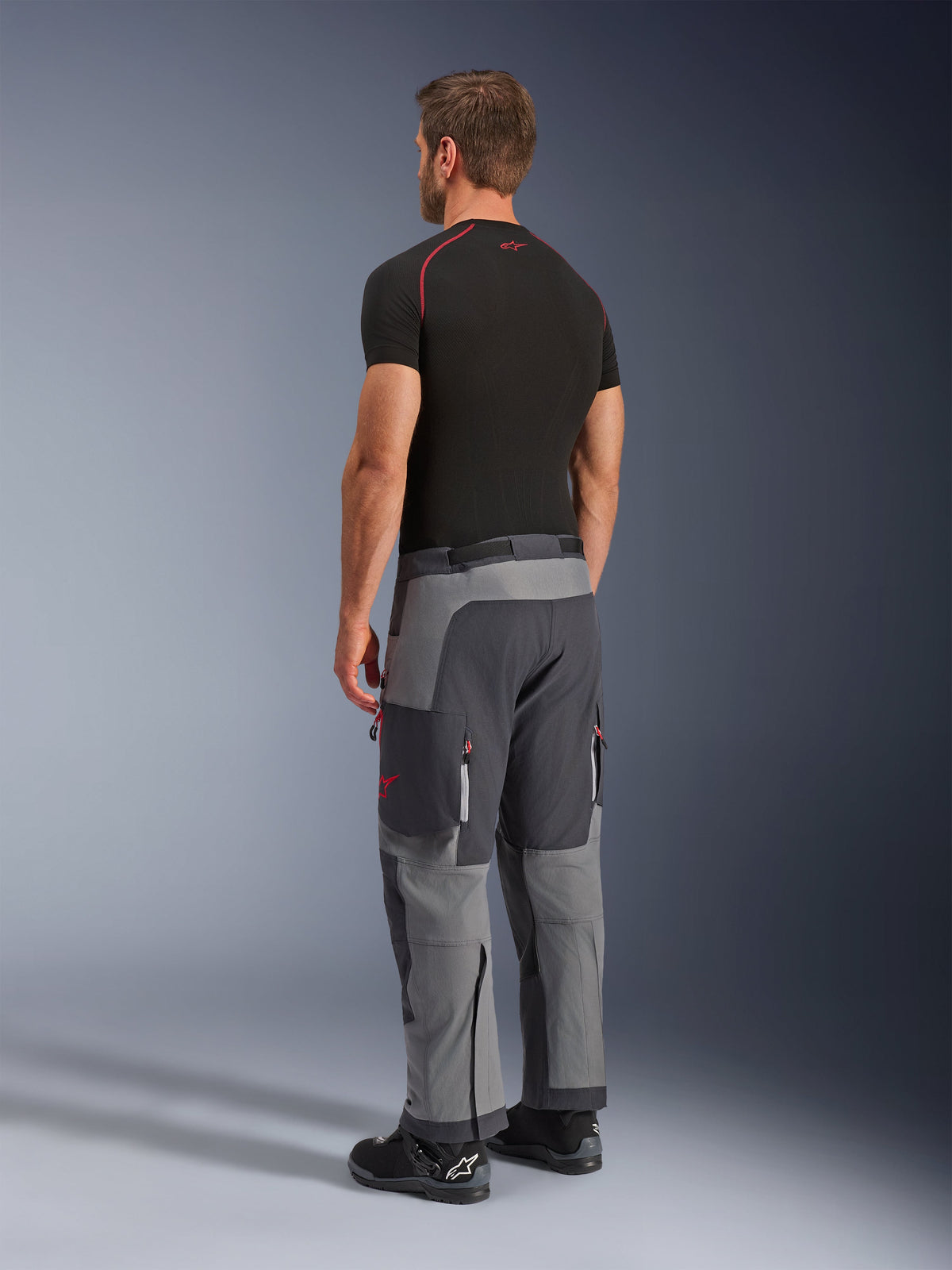 Alpinestars Maxdura Dual Pants, Adventure Motorcycle Pants, Dark Gray and Deep Gray, worn by a model from a back-side view, featuring red accents on zippers and logo, reinforced gray panels, cargo pockets, and over-the-boot fit