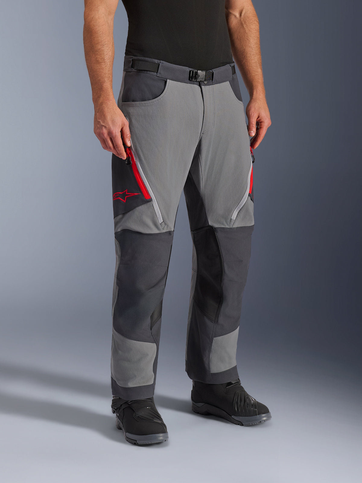 Alpinestars Maxdura Dual Pants in Dark Gray and Deep Gray, adventure-touring motorcycle pants featuring red zippered thigh vents and star logo, reinforced dark panels on knees, styled with black riding boots