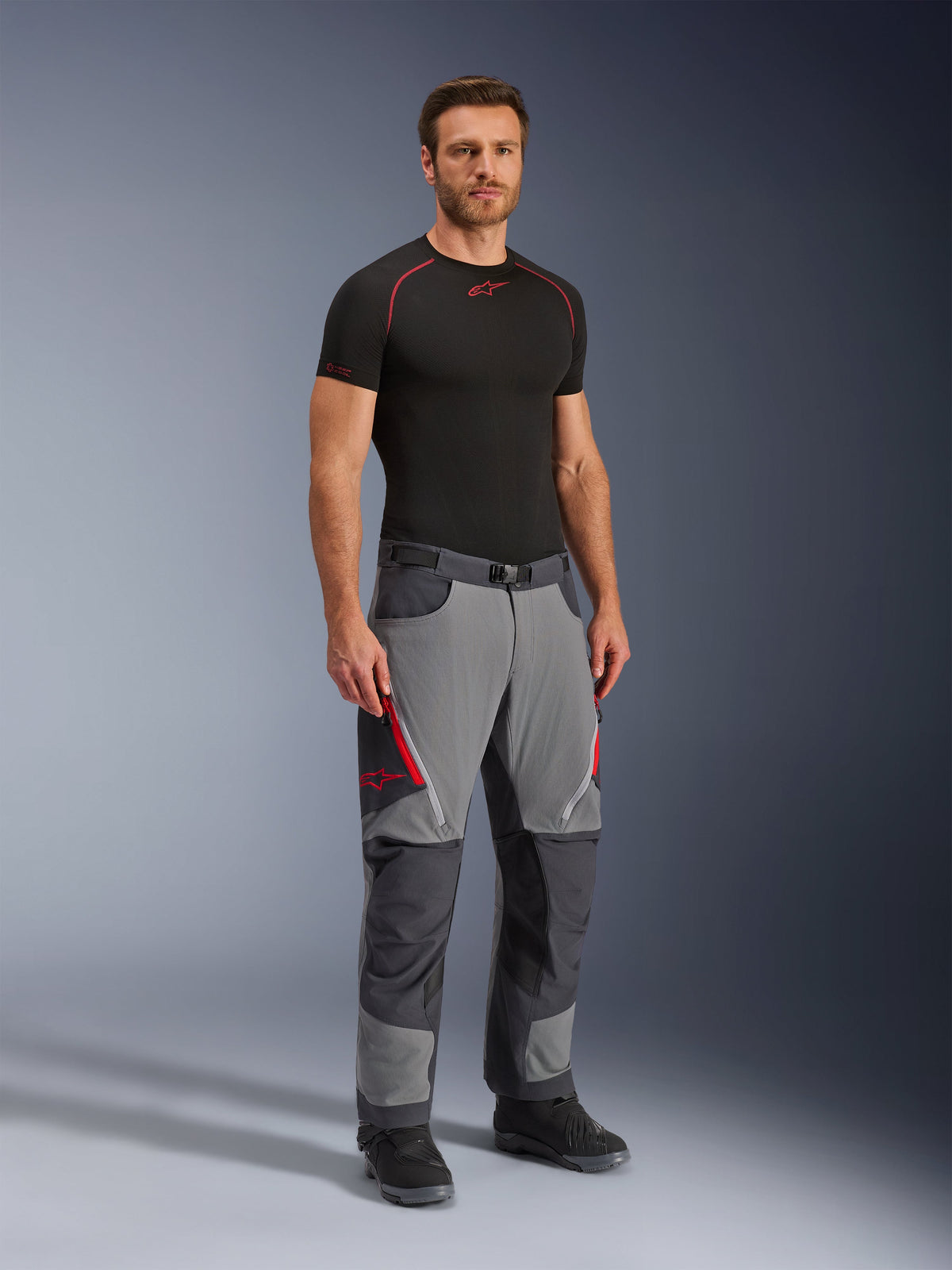 Alpinestars Maxdura Dual Pants, Adventure Motorcycle Pants, Dark Gray and Deep Gray, worn with a black base layer and boots, featuring red zippered vents, reinforced panels on knees and seat, and a belted waist, athletic fit for dual-sport riding