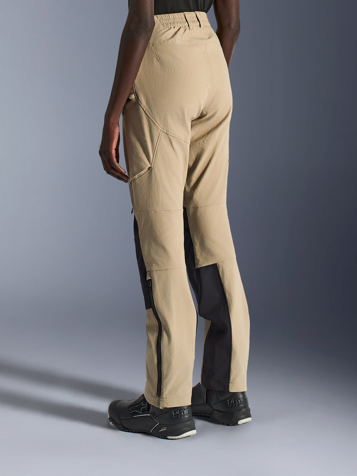 Alpinestars Woman Stella Flex-AST Explorer Pants in Sand and Black, rear and side view on a model, featuring lightweight ripstop construction, black reinforcement panels on the inner calves, zippered cargo pockets, and lower leg zippers for adventure riding.