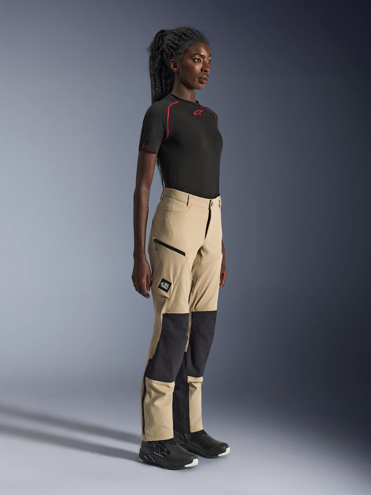 Alpinestars Woman Stella Flex-AST Explorer Pants, Sand and Black adventure touring pants, side profile on a model wearing a black Alpinestars t-shirt and riding boots, featuring ripstop knee reinforcements and zippered cargo pockets.
