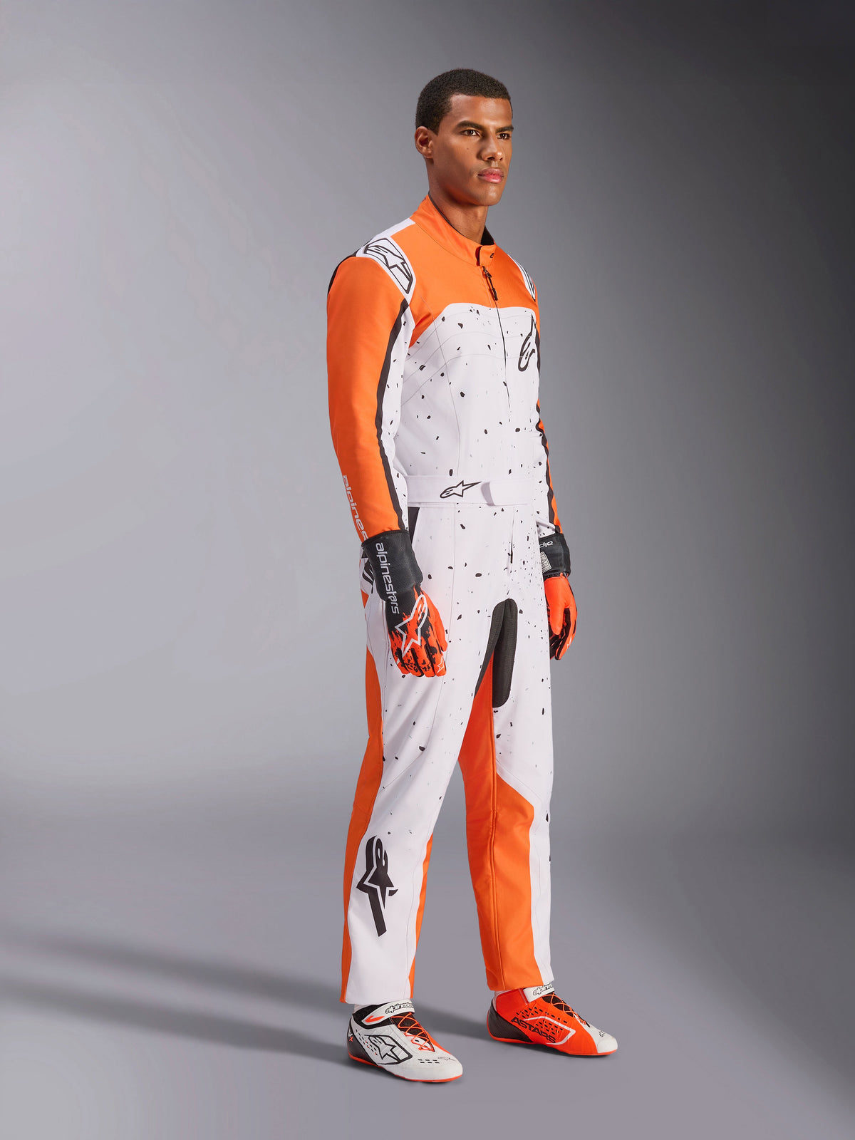 Alpinestars KMX-9 V3 Graphic 6 Karting Suit, White Orange and Black, featuring a speckled pattern and vibrant orange sleeves, worn by a model with matching racing gloves and shoes, professional dual-layer motorsport gear.