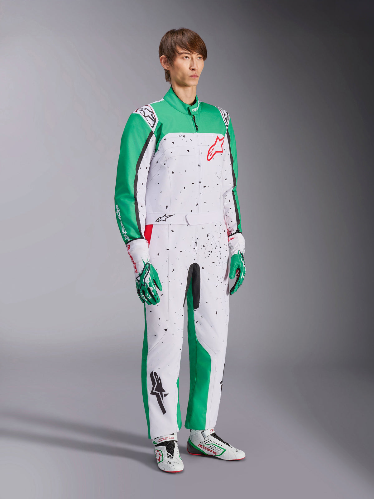 Alpinestars KMX-9 V3 Graphic 6 Suit, Karting Suit, White Green and Red, worn with matching racing gloves and shoes, features a white body with green sleeves and black splatter graphics, red Alpinestars logo on the chest, athletic one-piece design for motorsport.