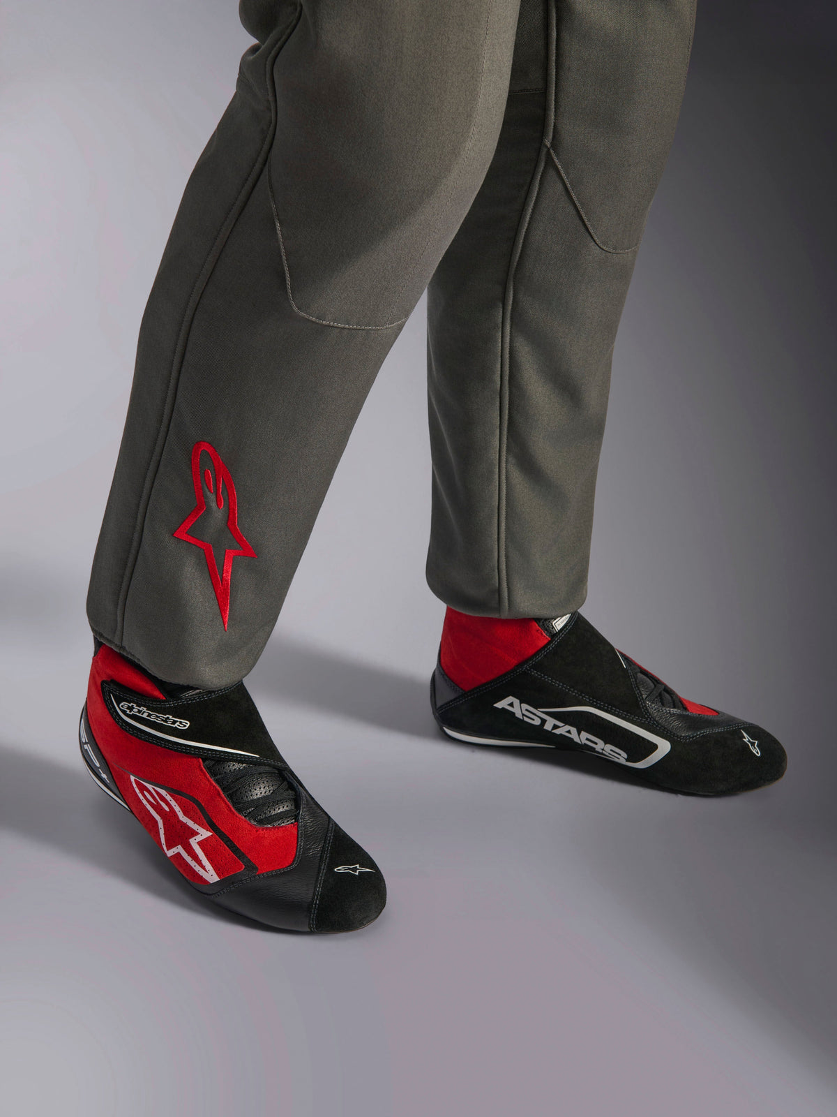 Alpinestars GP Race V3 Suit FIA, Anthracite Red racing suit, close-up of lower legs with embroidered red star logo, paired with red and black racing boots, professional automotive gear designed for driver protection and comfort