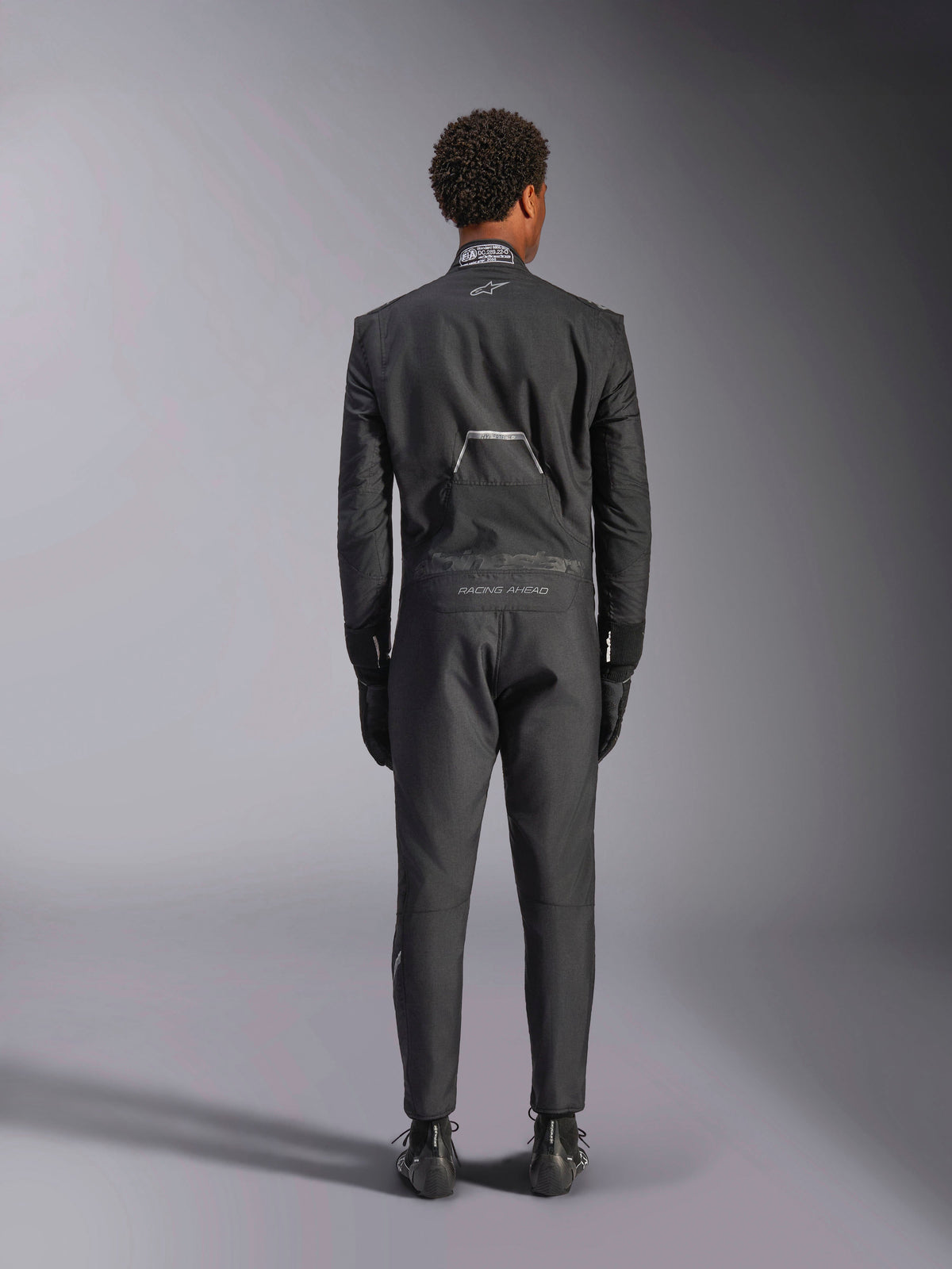 Alpinestars Hypertech V3 Suit, Black Dark Shadow, rear view of a professional one-piece auto racing suit, featuring FIA certification label on the collar and 'Alpinestars' and 'RACING AHEAD' branding on the lower back, worn with matching black racing gloves and boots.