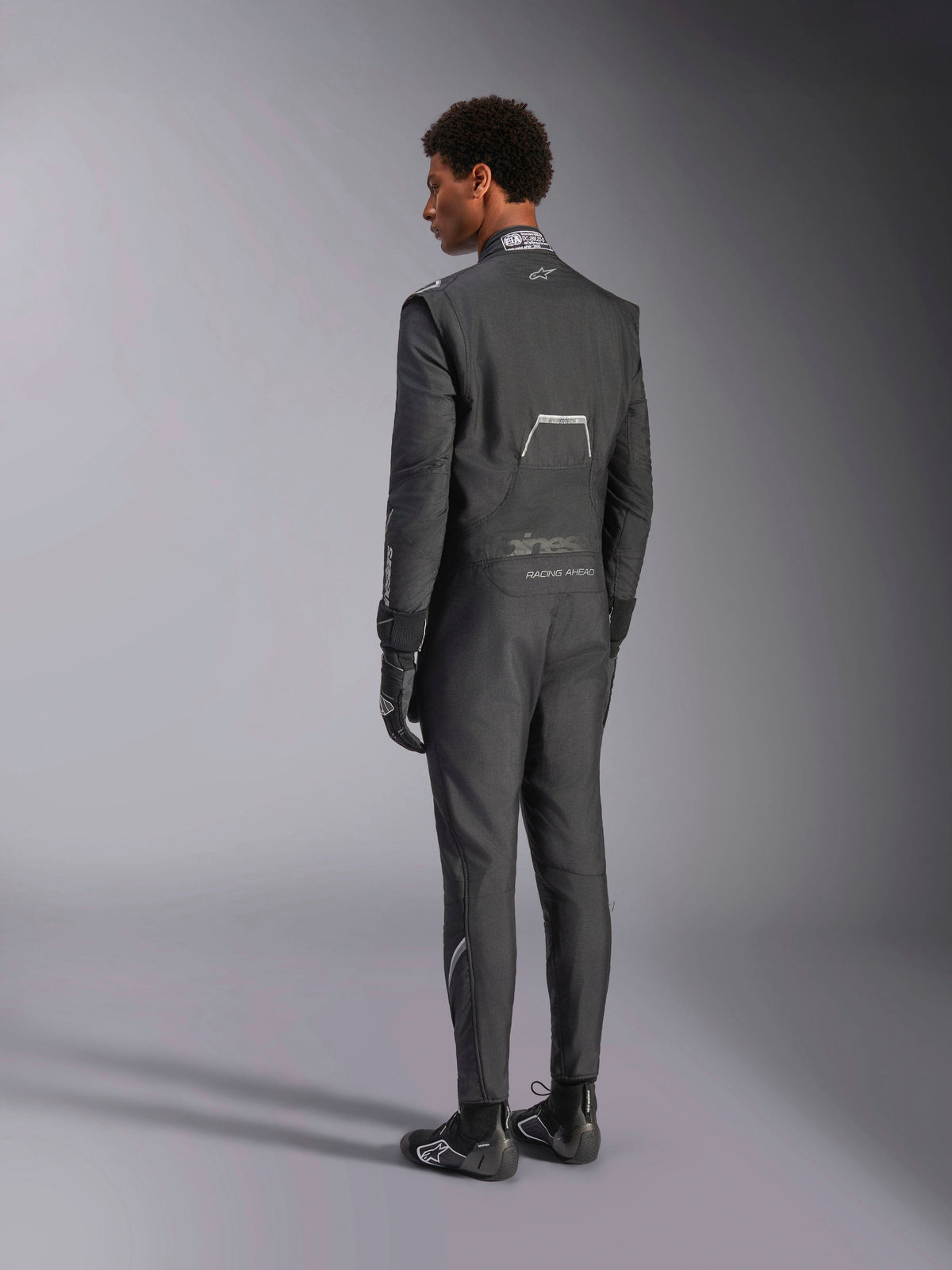 Alpinestars Hypertech V3 Suit, Black and Dark Shadow, rear view of male model wearing professional auto racing suit with matching black gloves and boots, featuring lightweight Nomex construction and FIA certification on the collar, athletic-slim fit for motorsport.