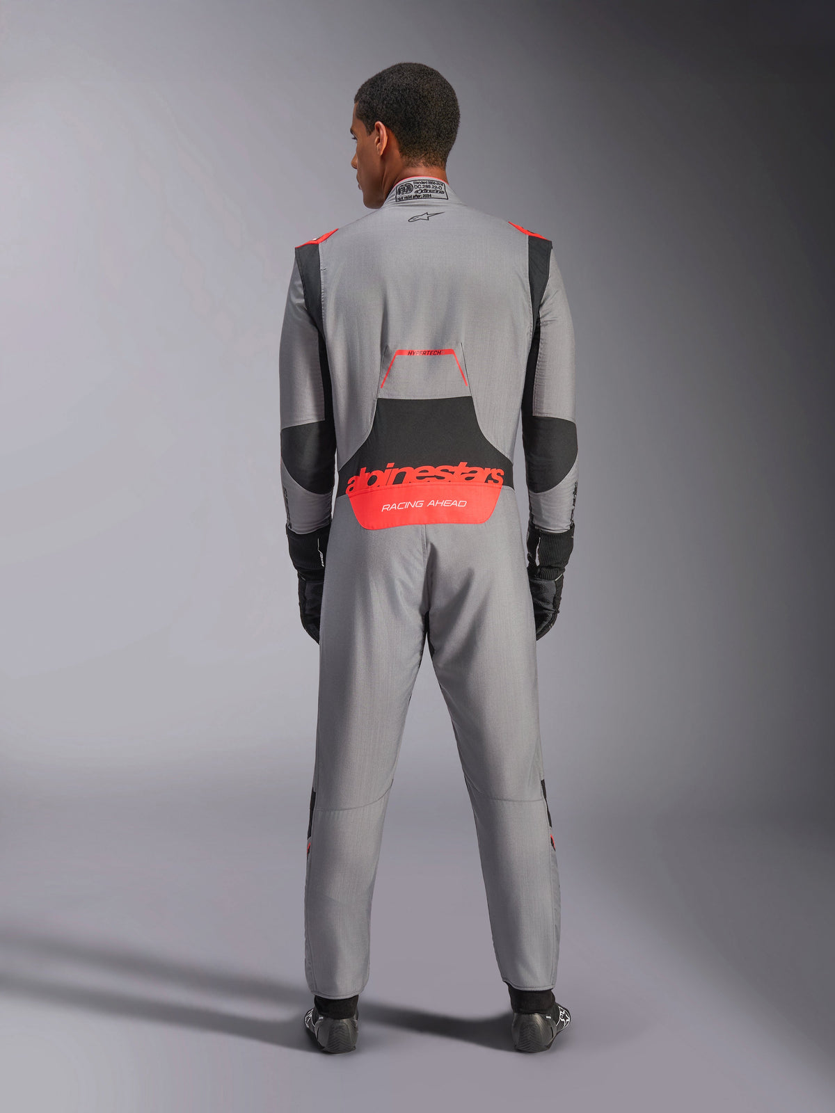 Alpinestars Hypertech V3 Suit, Professional Auto Racing Suit, Mid Gray with Black and Red Fluo accents, back view on a model featuring red Alpinestars branding on the lower back, lightweight Nomex construction for track performance.