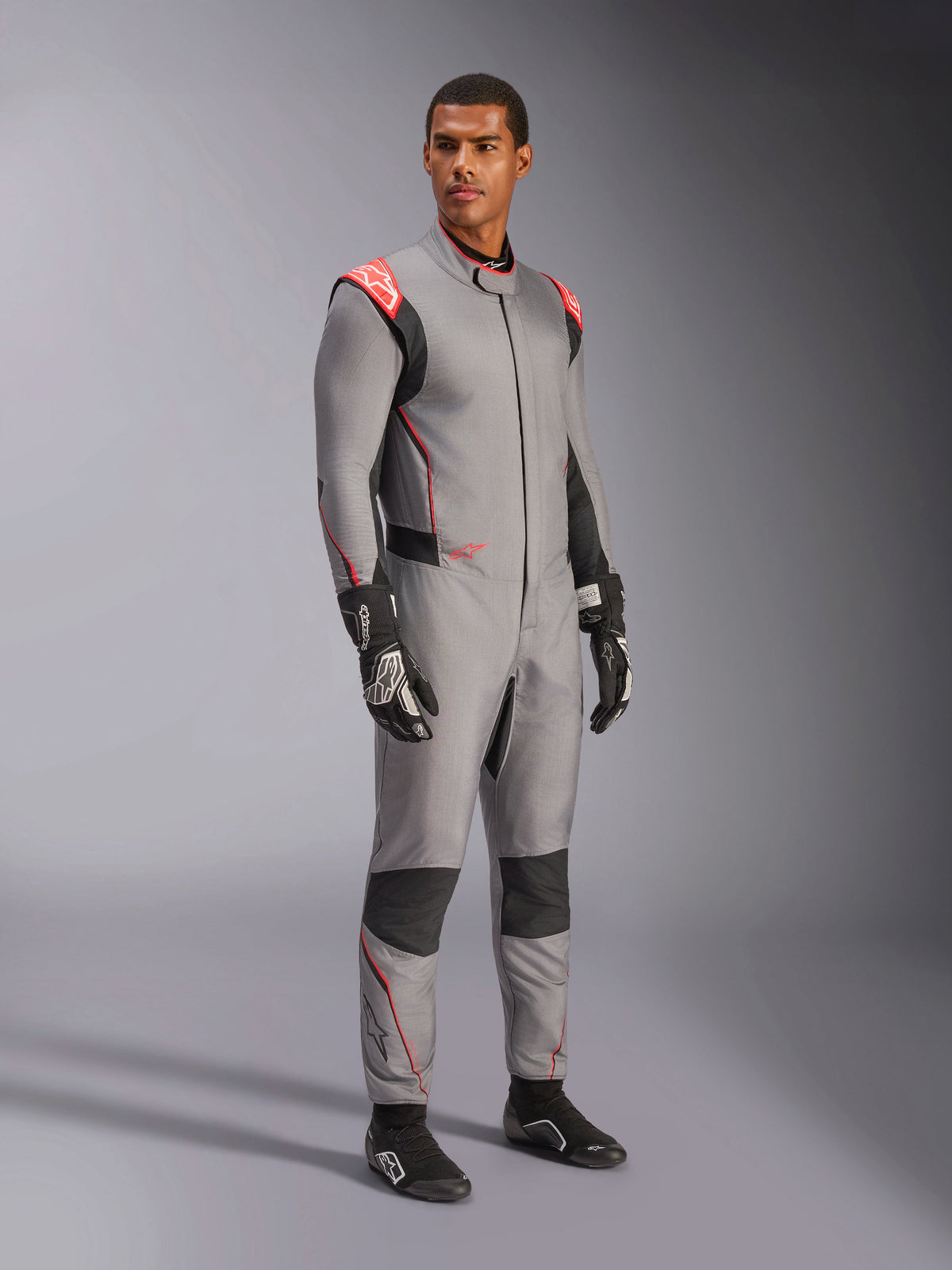 Male model wearing the Alpinestars Hypertech V3 Suit in Mid Gray with Black and Red Fluo accents, professional auto racing suit paired with black racing gloves and shoes, featuring a lightweight two-layer Nomex construction with red shoulder detailing and an athletic fit