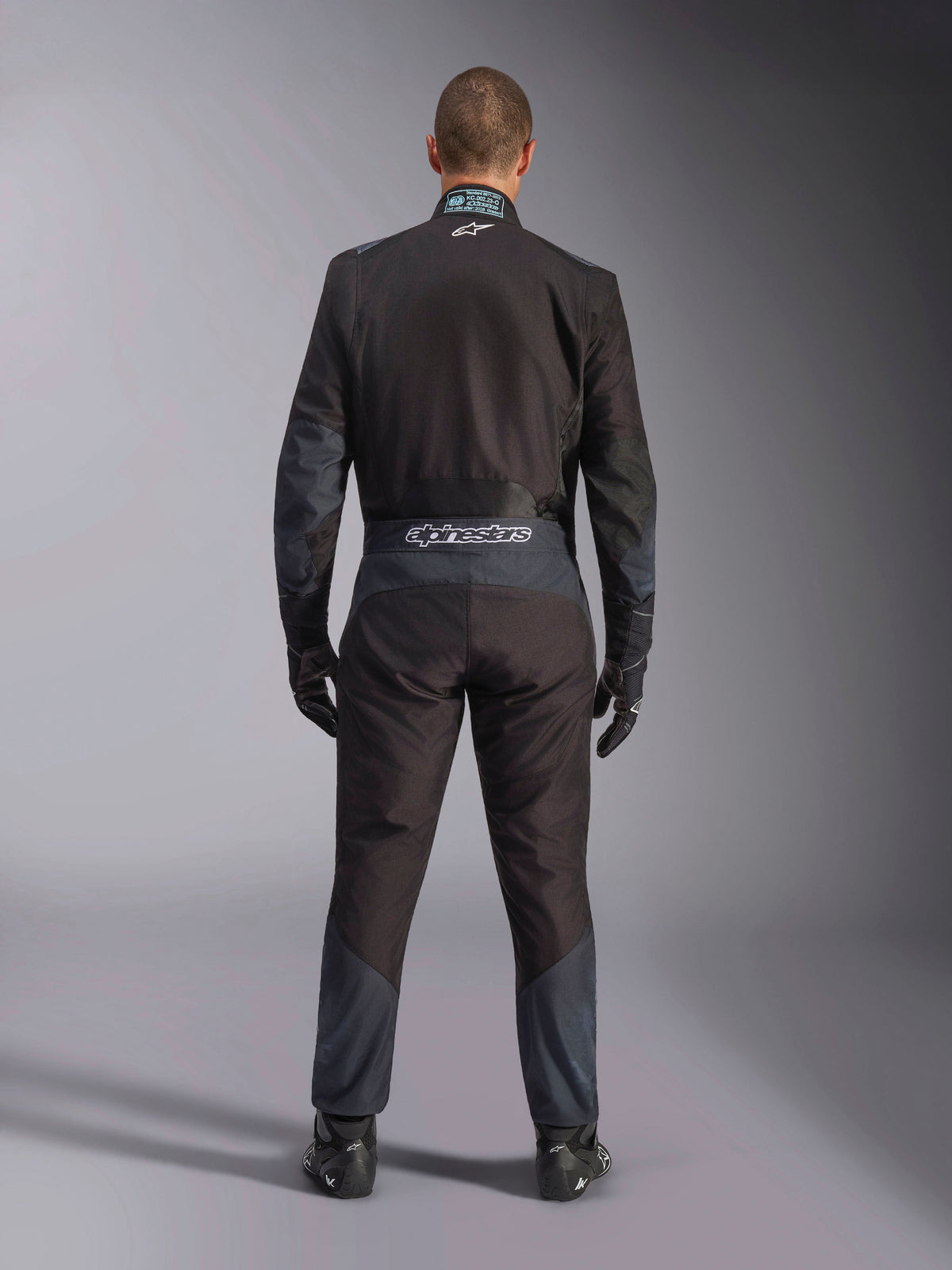 Alpinestars KMX-5 V3 Suit, Karting Suit, Black and Dark Shadow, back view of a driver wearing the full racing suit with matching gloves and shoes, featuring Alpinestars branding on the waistband and ergonomic paneling.