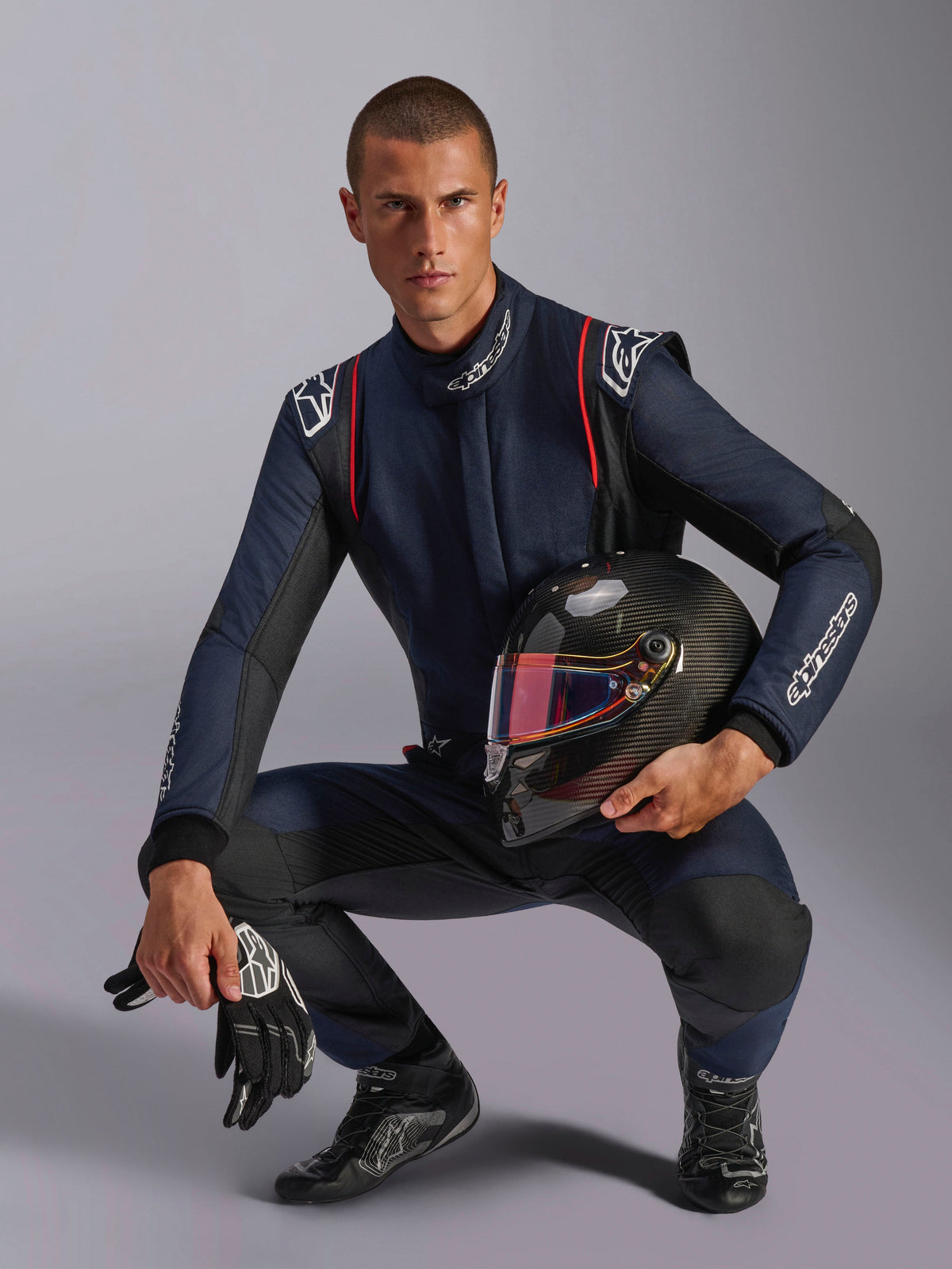 Male racing driver wearing Alpinestars GP Tech V4 Suit, Navy Blue with black and red accents, holding a carbon fiber helmet with an iridescent visor, black racing gloves and boots, professional automotive racing gear