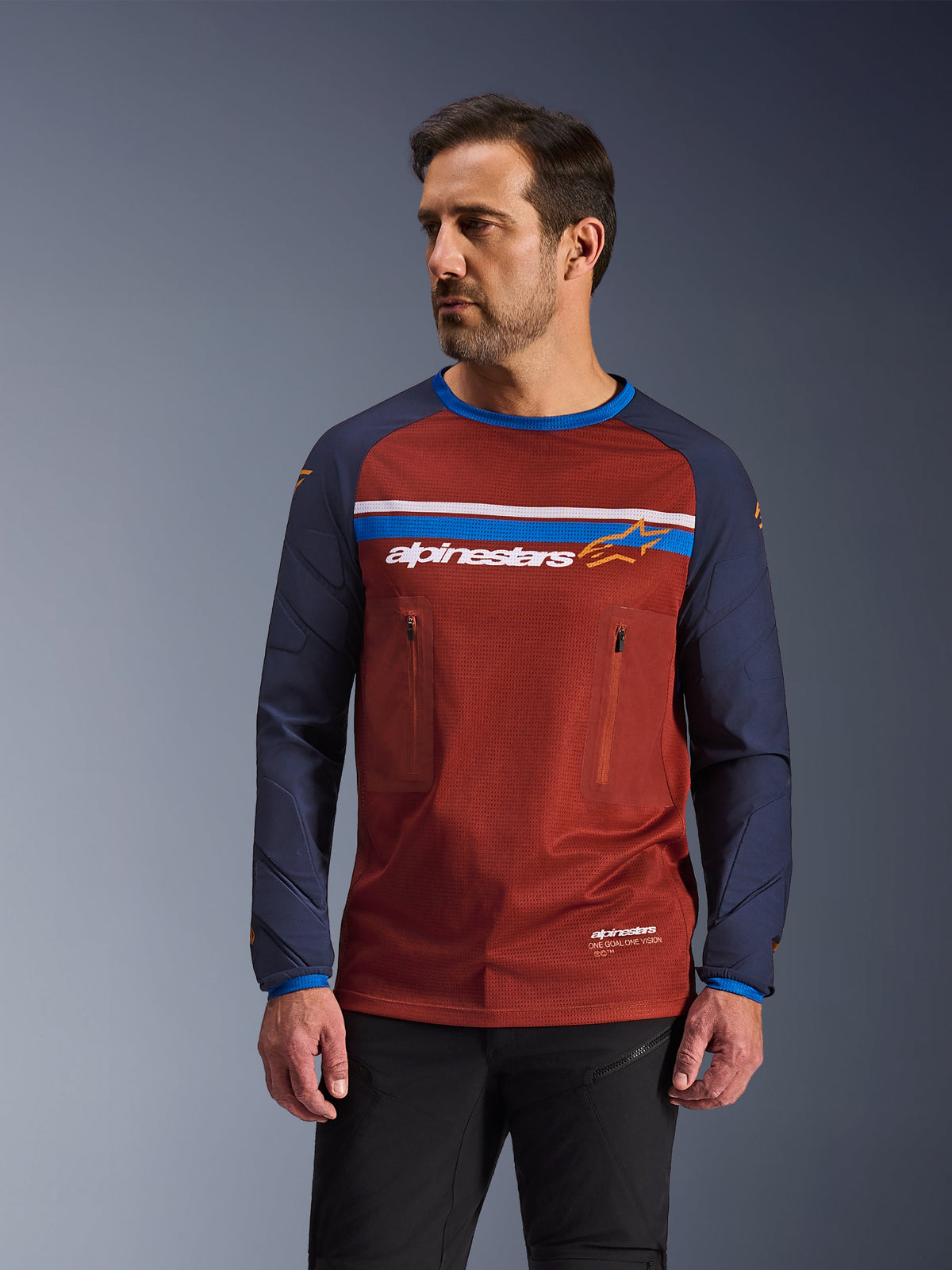 Male model wearing Alpinestars Maxdura Dual Jersey, Rust Brown and Navy Blue adventure motorcycle jersey, featuring dual zippered chest pockets, horizontal chest stripe with branding, technical mesh torso, and durable woven sleeves.