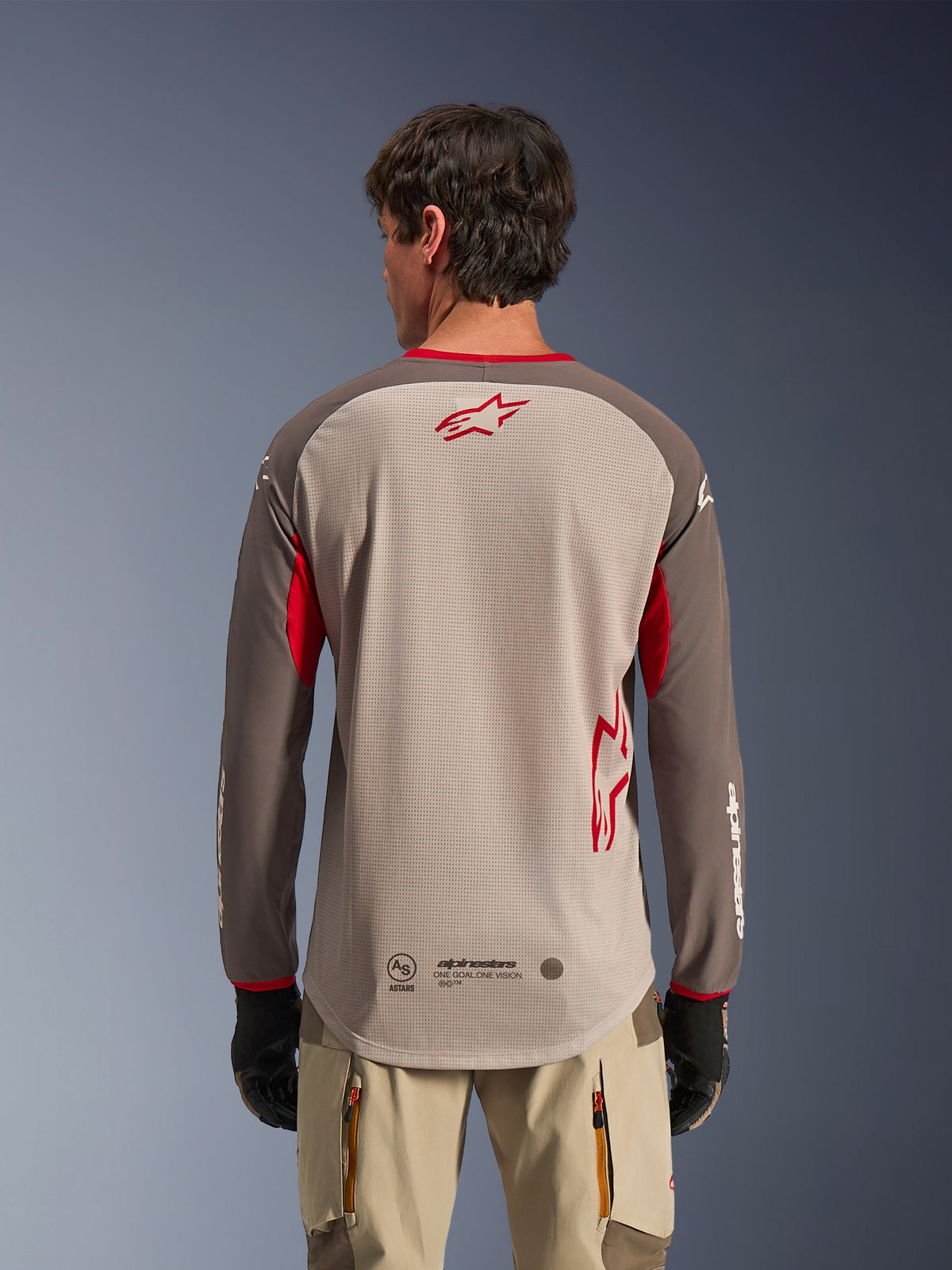Rear view of a man wearing the Alpinestars Maxdura Dual Jersey in Aluminum Walnut, adventure motorcycle jersey featuring a tan mesh torso with a red star logo, dark brown sleeves with red accents, paired with beige riding pants and black gloves, designed for high-performance off-road ventilation.