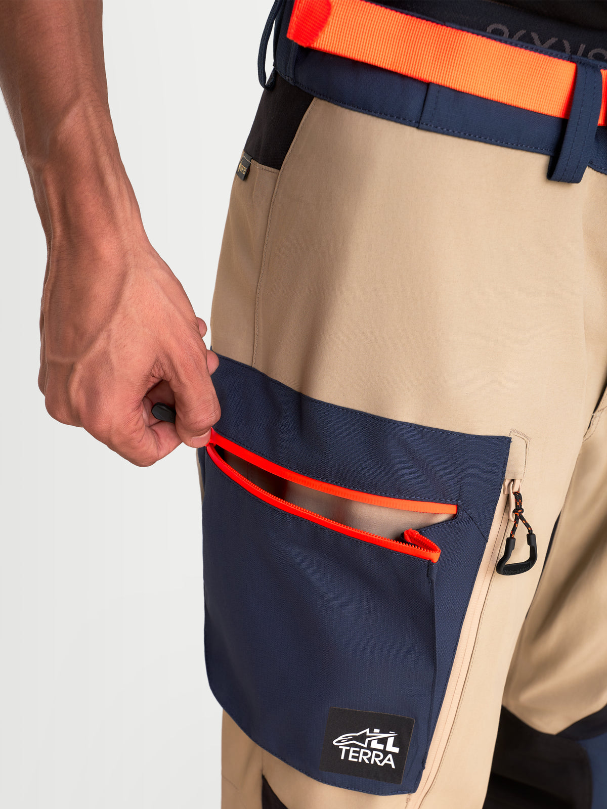 Alpinestars Arenis Gore-Tex Pants, men's adventure touring motorcycle pants in beige and navy blue, close-up view showing zippered cargo pocket and waist adjustment system worn by a model.