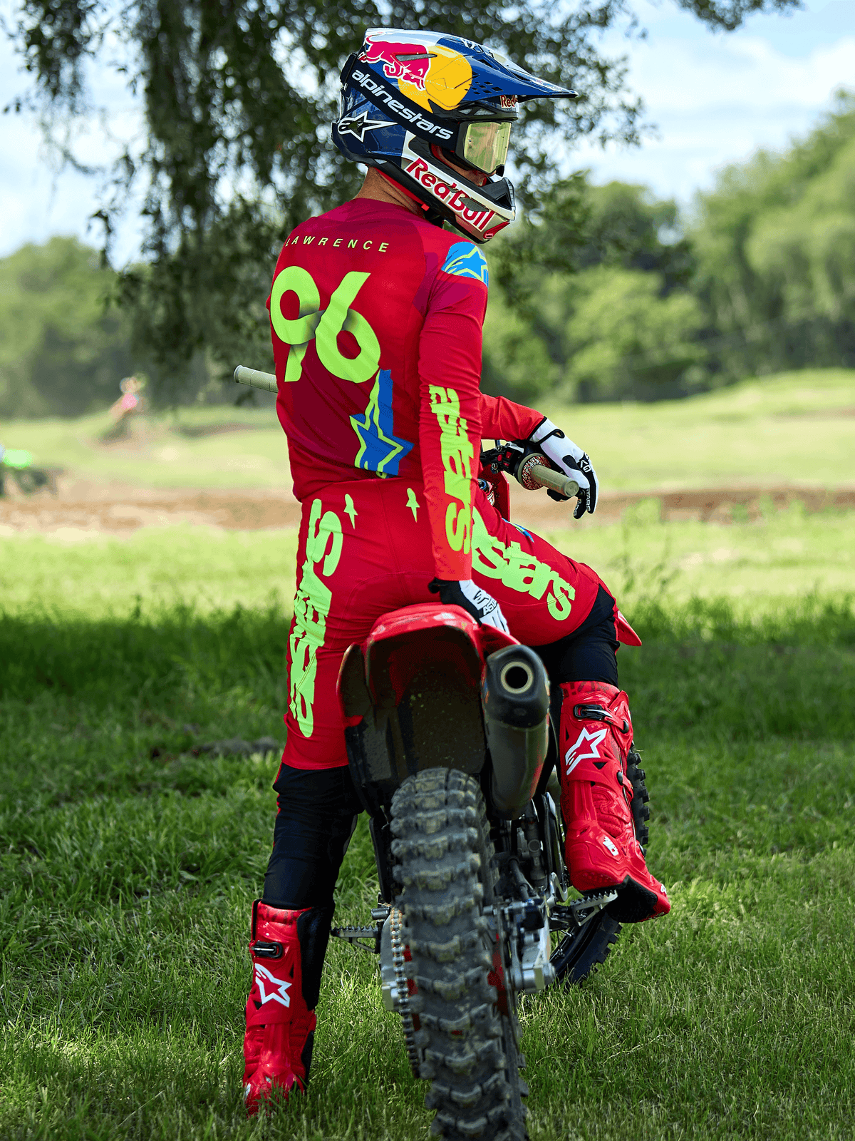Motocross rider sitting on a red dirt bike, Alpinestars 2025 Supertech Maker Pants in Bright Red and Yellow Fluo, matching red jersey with '96' and 'LAWRENCE' on the back, red racing boots, and a Red Bull branded helmet, professional off-road racing gear set