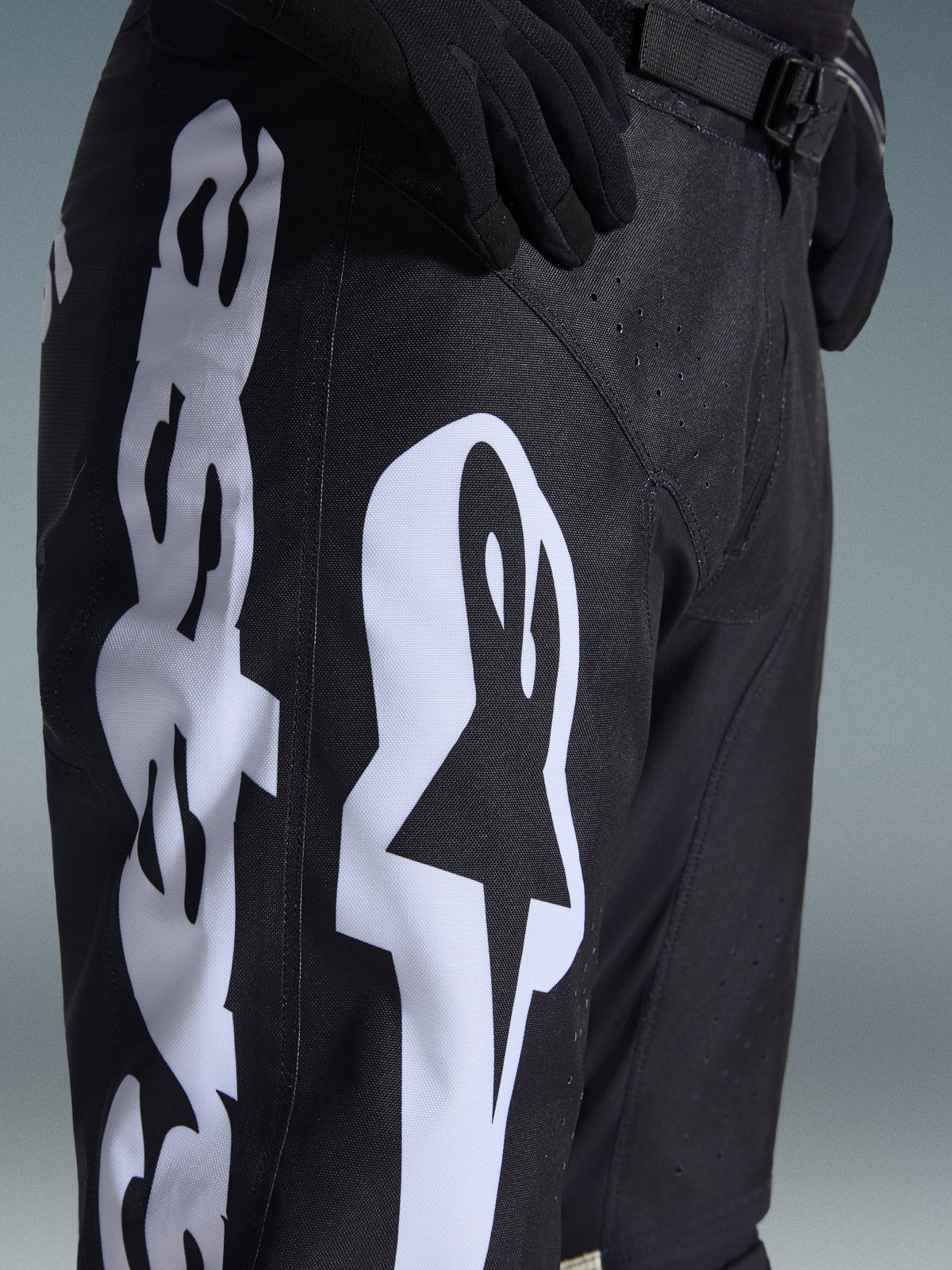 Alpinestars 2026 Racer Air Riway Pants, black motocross pants with bold white Alpinestars logo on the thigh, close-up showing technical fabric and adjustable waist strap, worn with black gloves for off-road riding