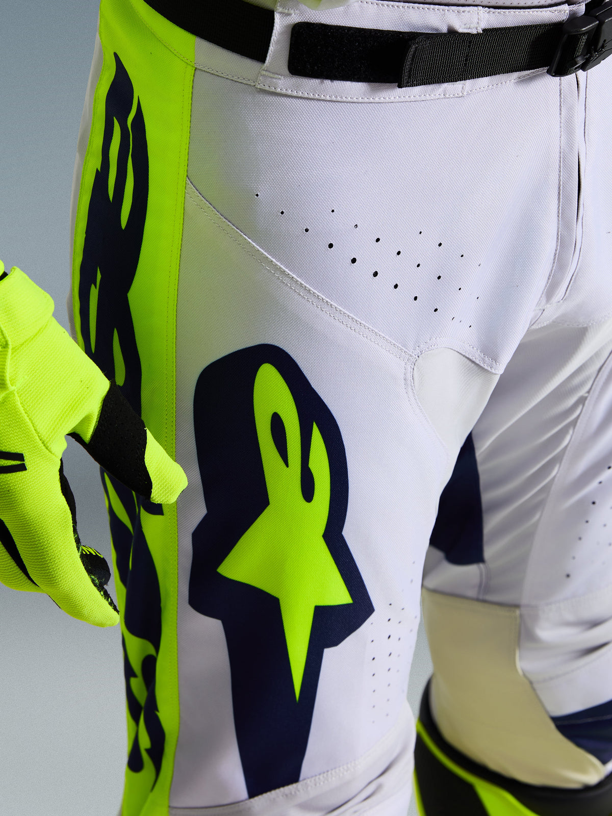 Alpinestars 2026 Racer Air Riway Pants, Close-up of white and navy motocross pants with vibrant yellow panels, featuring laser-cut ventilation holes for airflow and bold Astar branding, paired with matching yellow glove