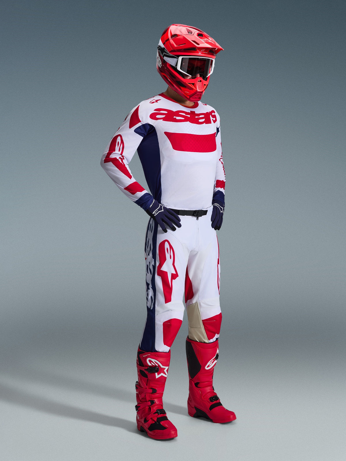 Motocross rider wearing full racing gear, Alpinestars 2026 Racer Riway Pants in White, Blue and Red, featuring a white base with bold red logos and blue side panels, paired with a matching white and red jersey, red helmet, and red motocross boots.