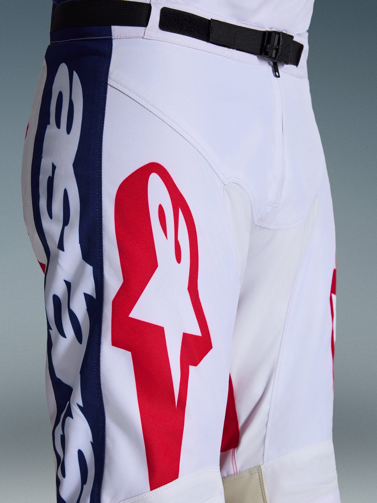 Alpinestars 2026 Racer Riway Pants, Motocross Pants, White Blue Red, close-up of the upper leg and waist showing large red star logo on white polyfabric, blue side panels with branding, and black adjustable waist belt with SX2 magnetic buckle, sporty off-road racing design