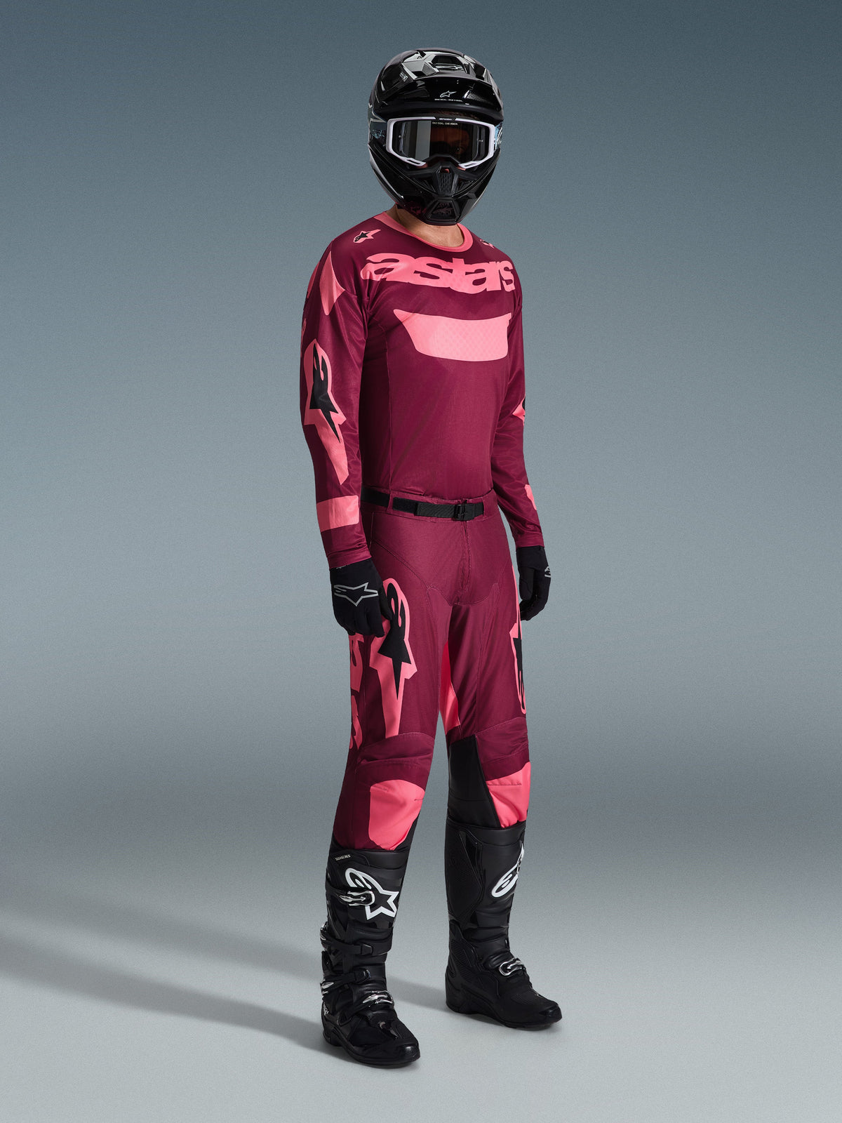 Motocross rider wearing complete racing gear, Alpinestars 2026 Racer Riway Jersey in Dark Red Coral with pink accents, matching dark red pants with pink logos, black helmet with white-rimmed goggles, black gloves, and black boots with white logos, coordinated outfit for off-road racing