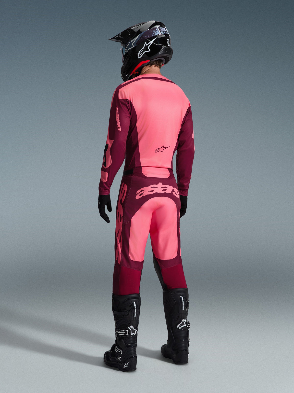 Alpinestars 2026 Racer Riway Jersey and matching pants, Motocross gear in Dark Red Coral, rear view of rider wearing black helmet and black boots, featuring vibrant coral and maroon panels with bold Astars branding on the back