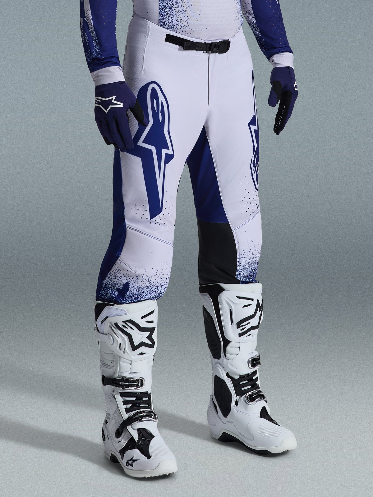 Alpinestars 2026 Supertech Scenz Pants, Motocross Pants, Light Gray and Dark Blue, worn with white motocross boots and blue gloves, featuring large blue Alpinestars logos on the thighs, black knee panels, and a stippled graphic design, professional off-road racing gear