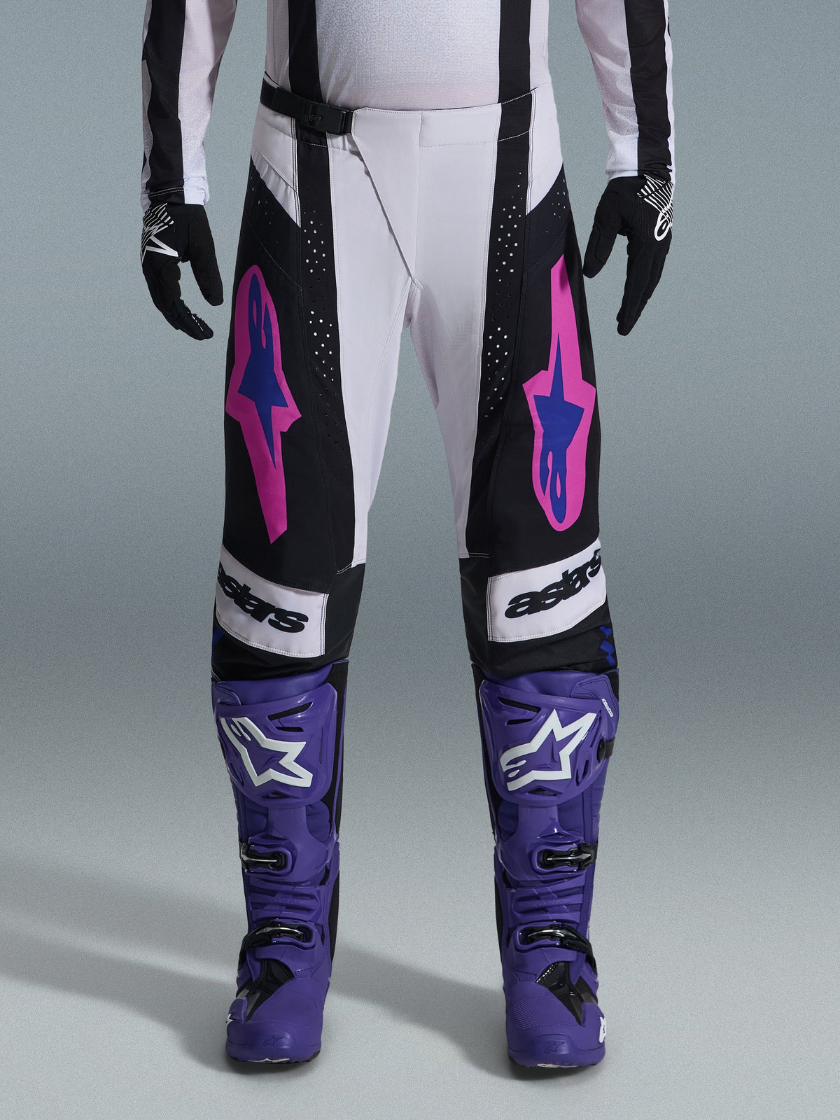 Motocross rider wearing the Alpinestars 2026 Techstar Knif Gear Set, white and black pants with pink and blue logos, purple boots with white branding, black gloves, featuring an athletic race fit and laser-cut ventilation for off-road racing