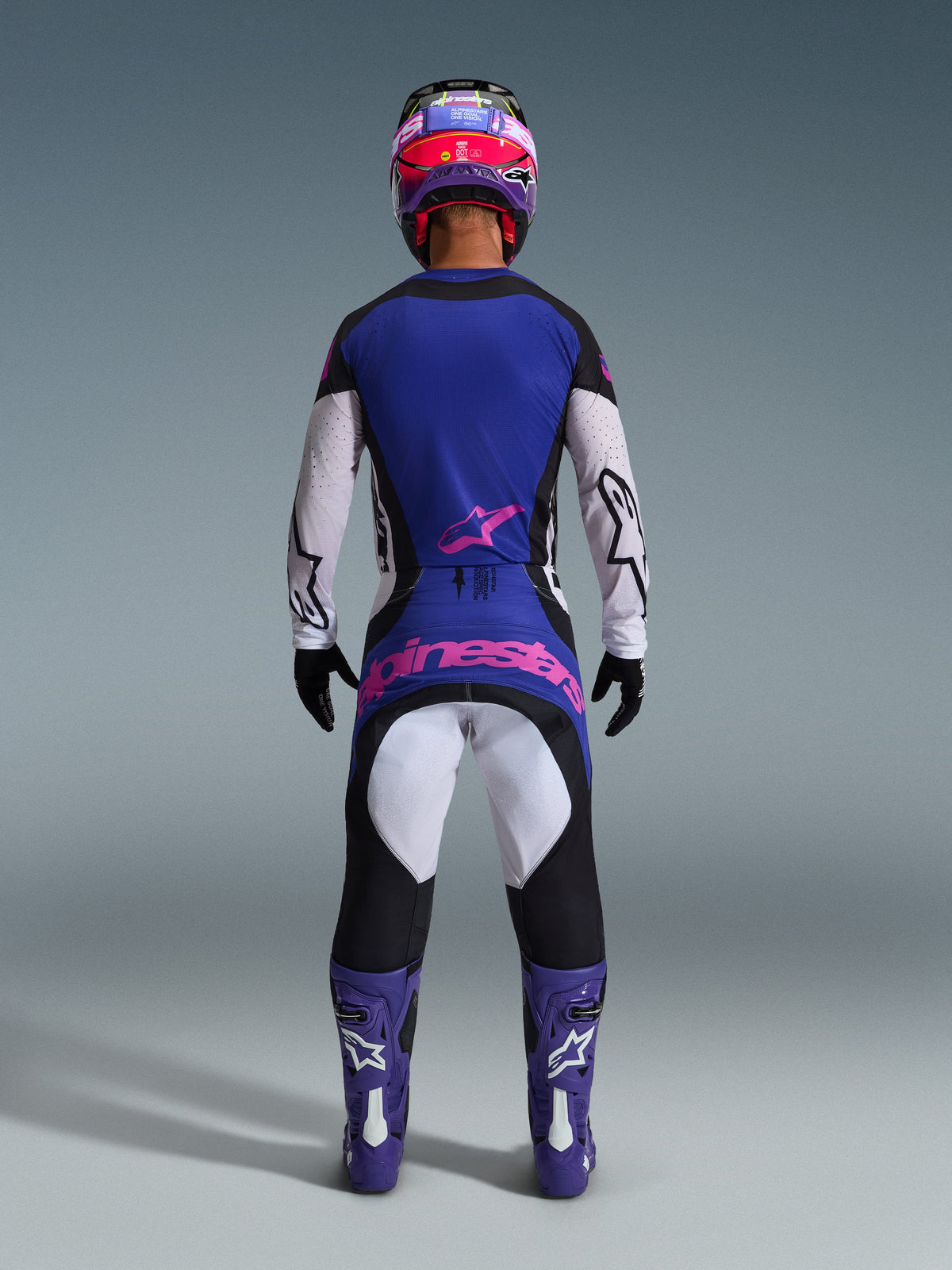 Rear view of a motocross rider wearing the Alpinestars 2026 Techstar Knif Gear Set, blue and white jersey with a pink star logo, matching blue and black pants featuring large pink Alpinestars branding on the rear, purple and white boots, and a purple and red helmet, coordinated professional off-road racing outfit