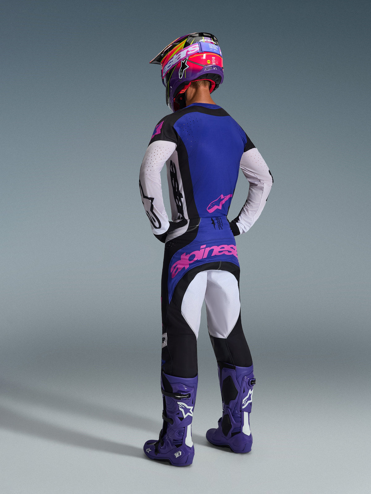 Motocross rider seen from the back wearing the Alpinestars 2026 Techstar Knif Gear Set, blue jersey with white perforated sleeves and pink logos, black and white pants with pink branding on the rear, paired with purple boots and a vibrant purple and pink helmet, professional motocross racing outfit.