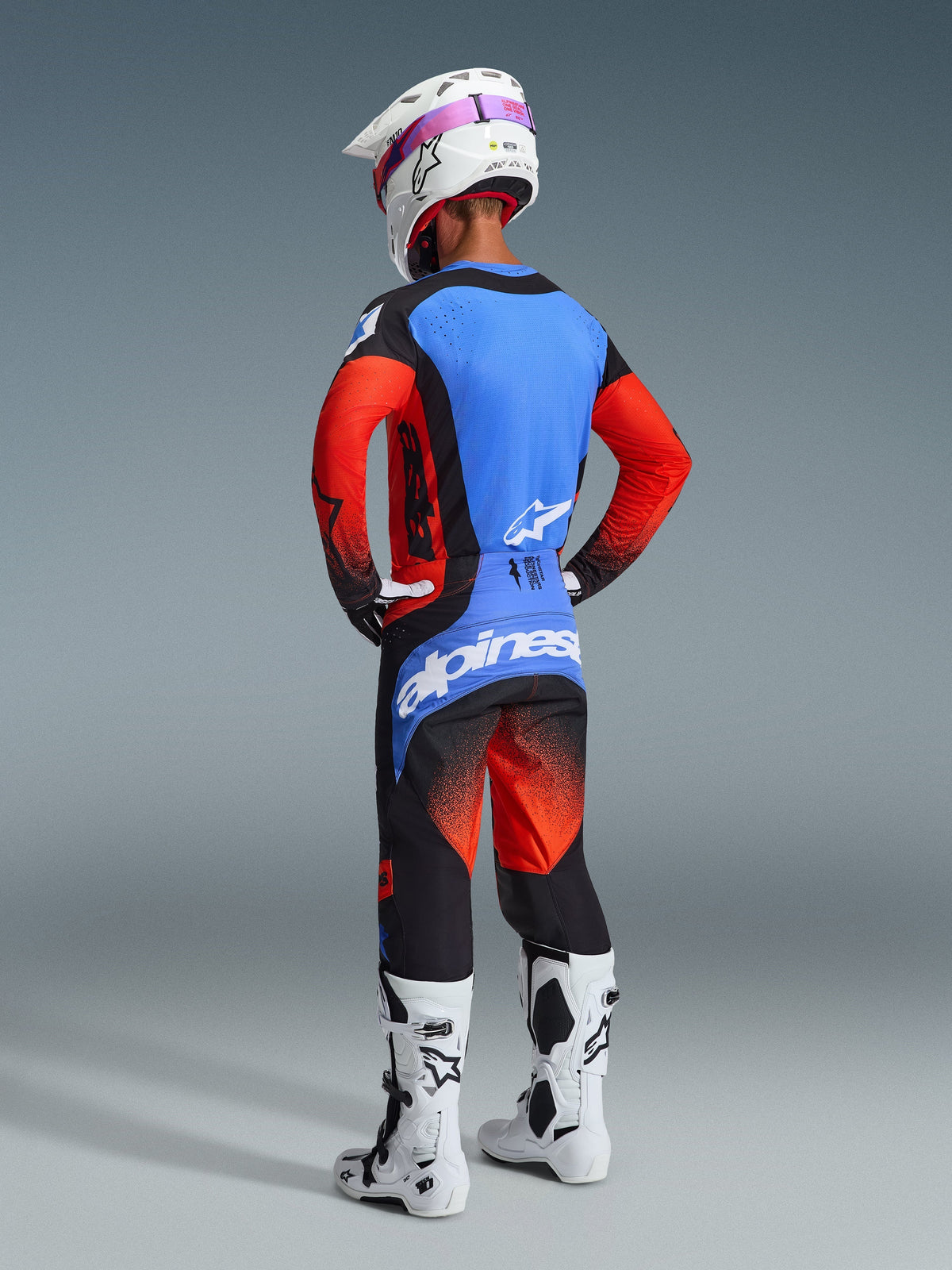 Motocross rider from behind wearing complete racing gear, 2026 Techstar Knif Pants in Orange Black and UCLA Blue, blue jersey with orange sleeves, white helmet with purple goggles, white and black boots, coordinated outfit for off-road performance