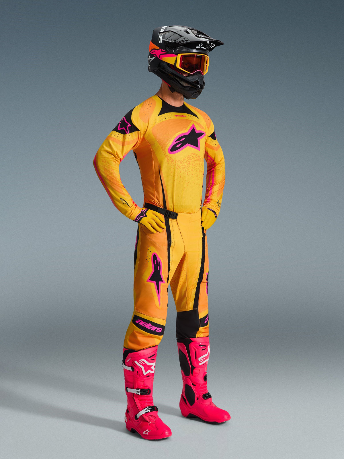 Motocross rider wearing Alpinestars 2026 Techstar Nomur Pants in Orange Yellow Pink, complete racing gear set with matching jersey and gloves, bright pink boots, black helmet with orange goggles, full body view against a grey background, professional off-road racing outfit