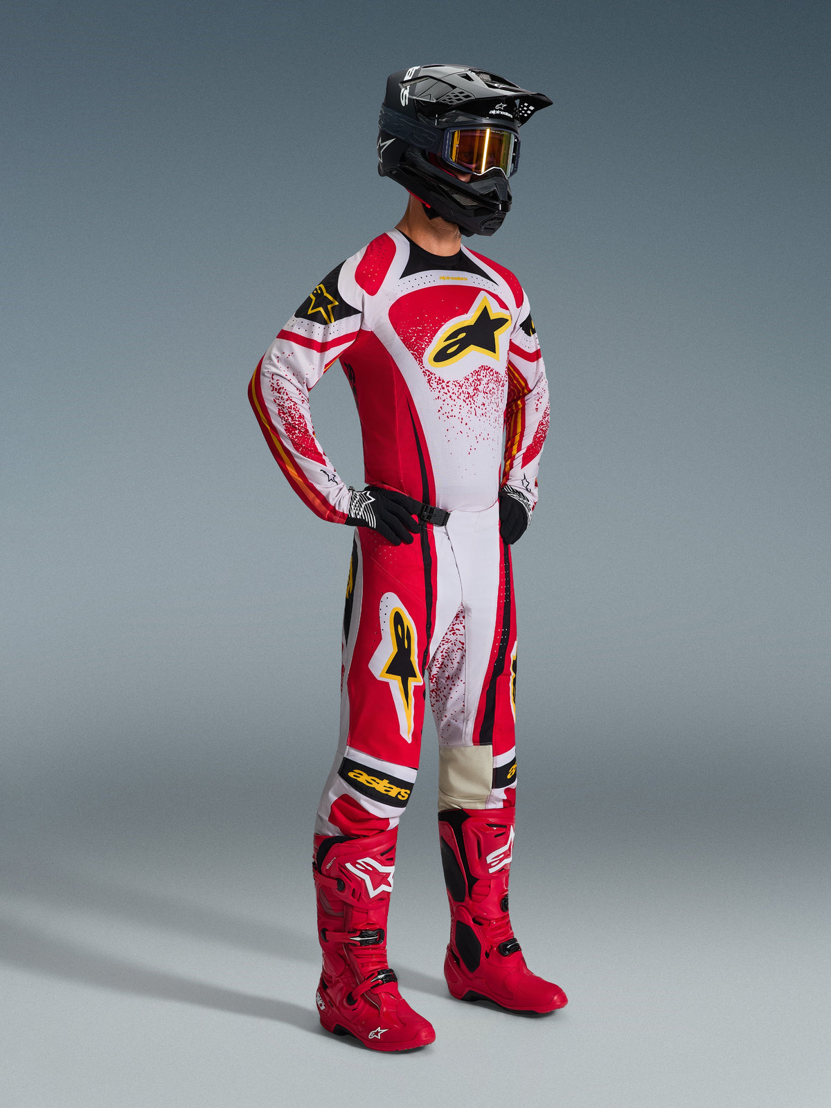 Motocross rider wearing the Alpinestars 2026 Techstar Nomur Gear Set, Red and White with Black accents and yellow logos, worn with matching red boots and a black helmet, professional racing outfit for off-road competition
