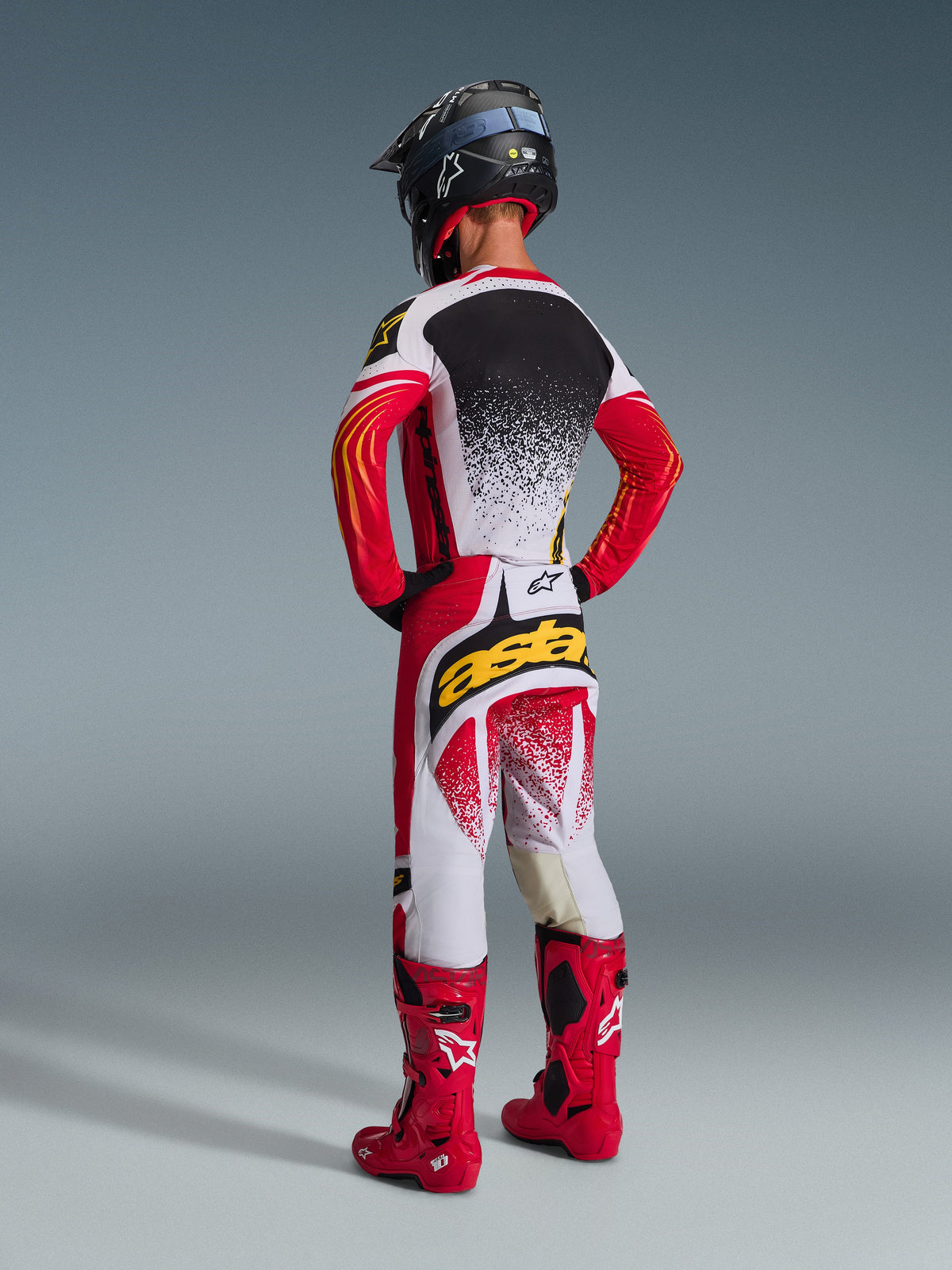Motocross rider from behind wearing the Alpinestars 2026 Techstar Nomur Gear Set, Red white and black jersey with a white speckled back, matching pants with yellow 'astars' logo on the seat, bright red racing boots and a black helmet, complete coordinated motocross outfit